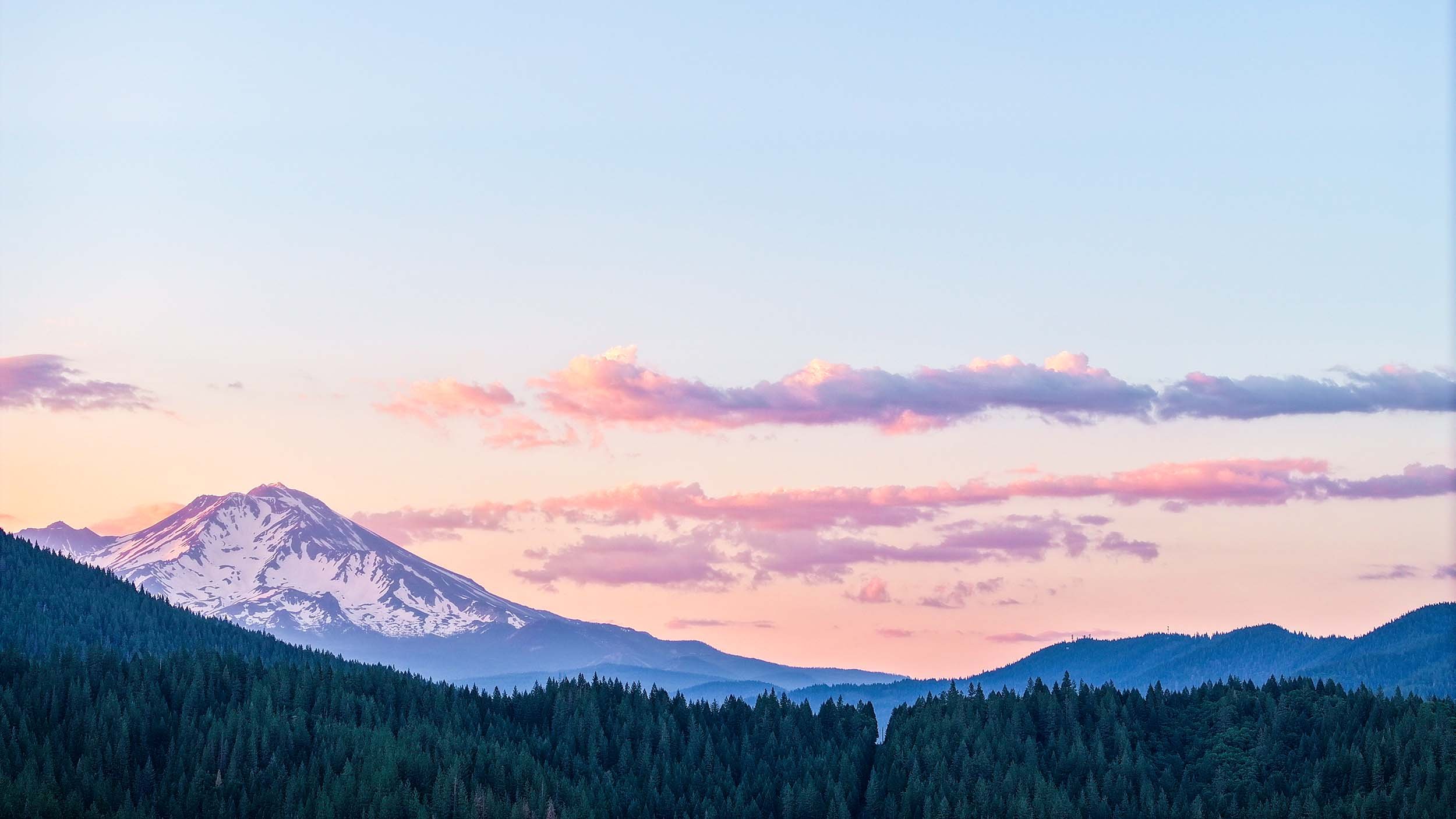 Snow-capped mountain with forested slopes and pink clouds at dusk.