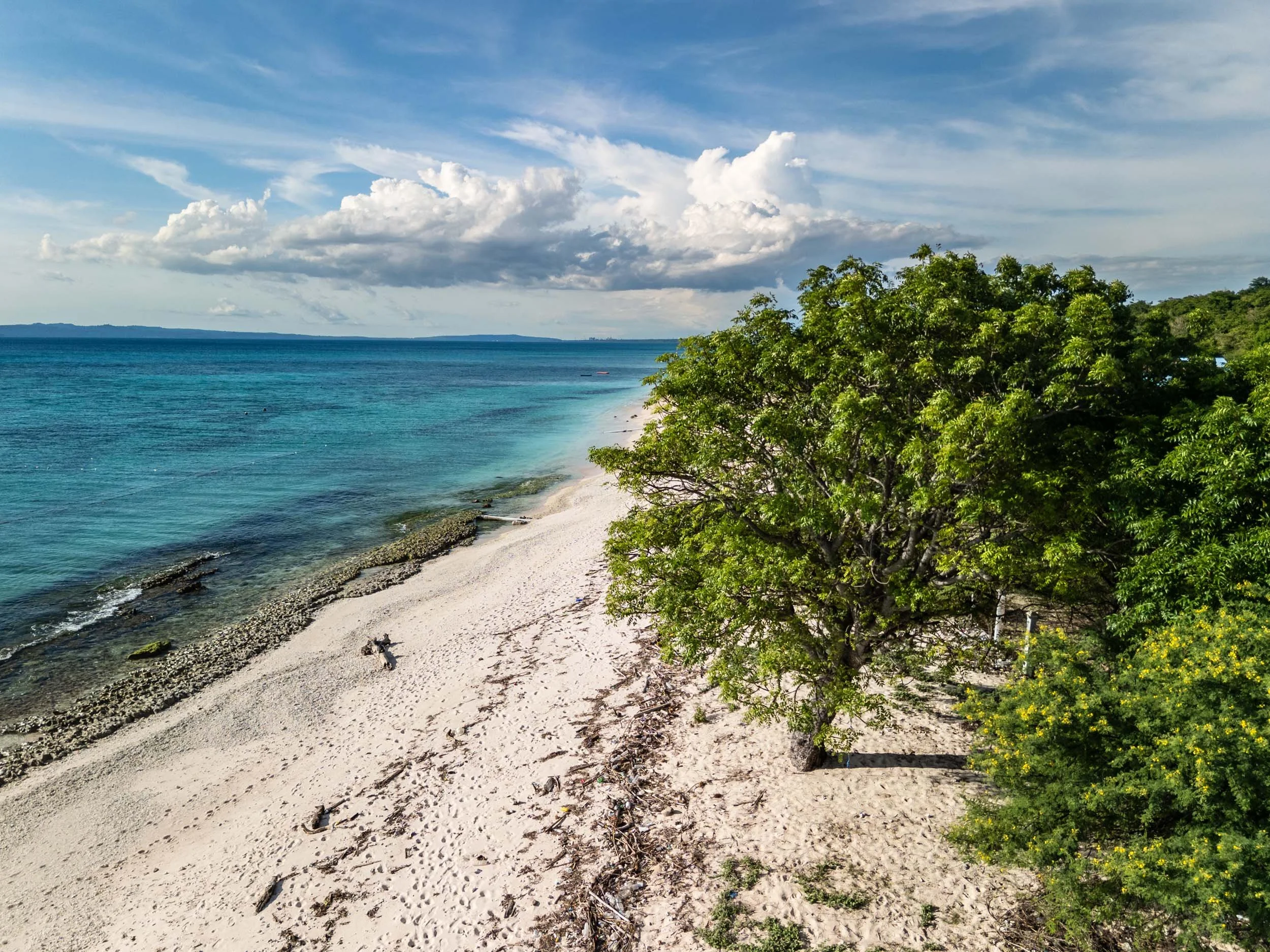 A sandy beach with clear turquoise water, green trees on the right, and a partly cloudy sky.
