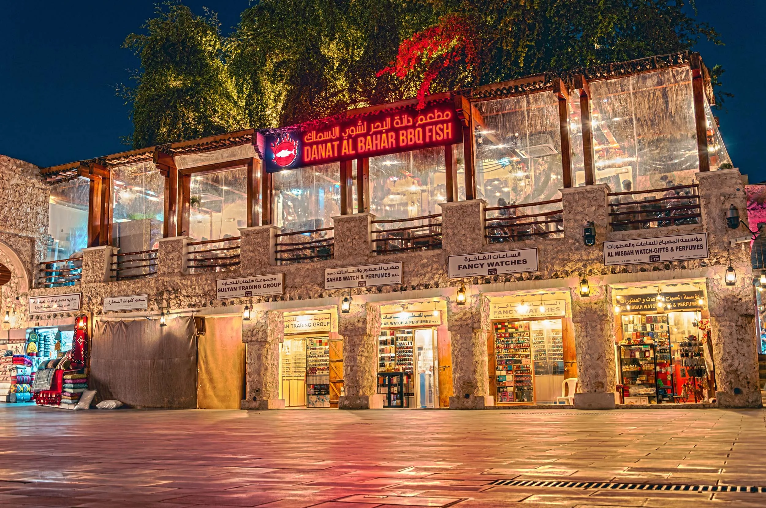 Night view of a two-story stone building with a red neon sign in Arabic and English reading 'DANAT AL BAHAR BBQ FISH.' The upper level has a glass-enclosed balcony with trees behind it. The lower level features stores with illuminated displays, selli