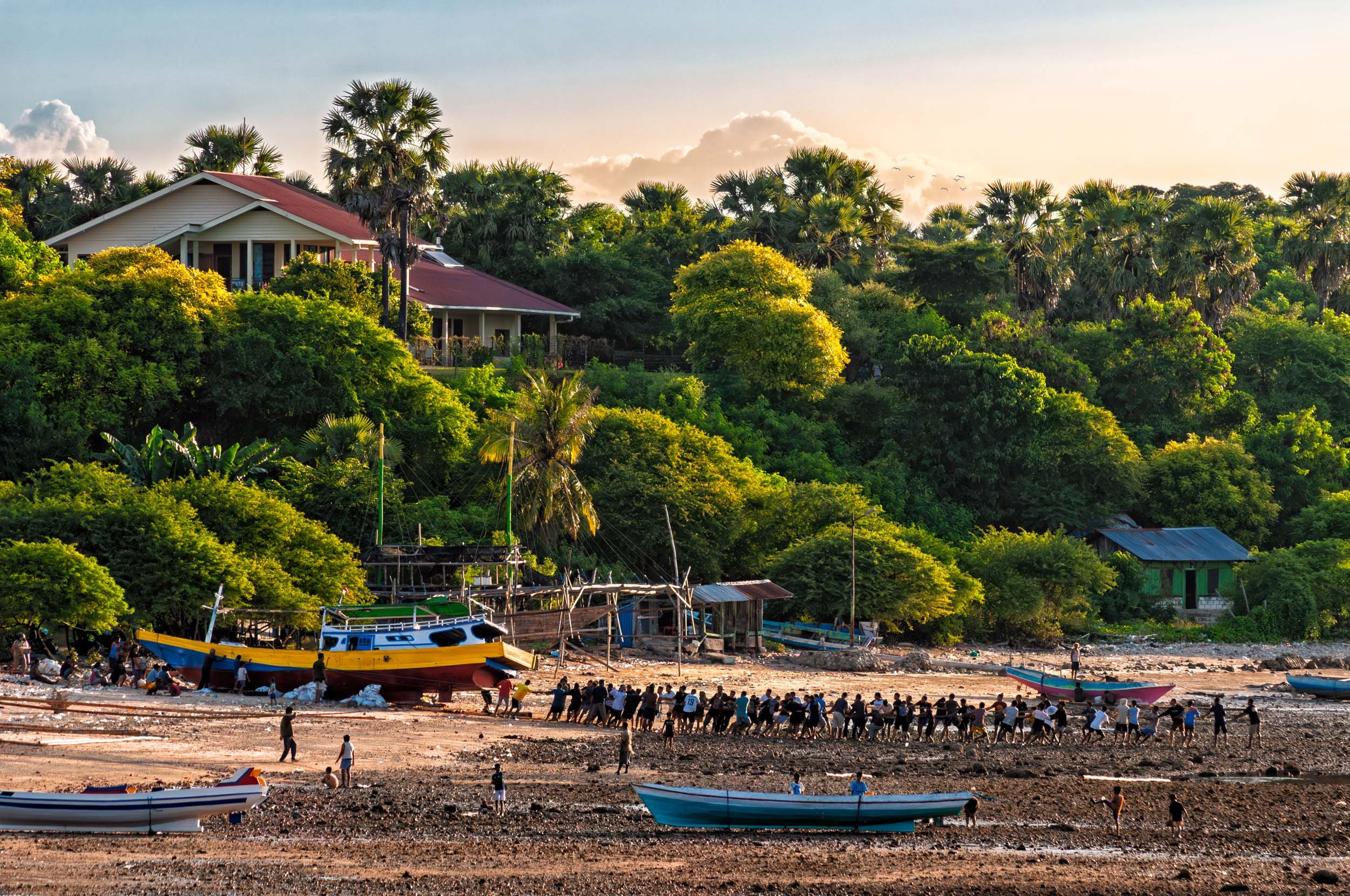 A coastal scene showing a group of people pulling a boat into the sea, several small boats are nearby, lush green foliage and houses on a hillside during sunset.