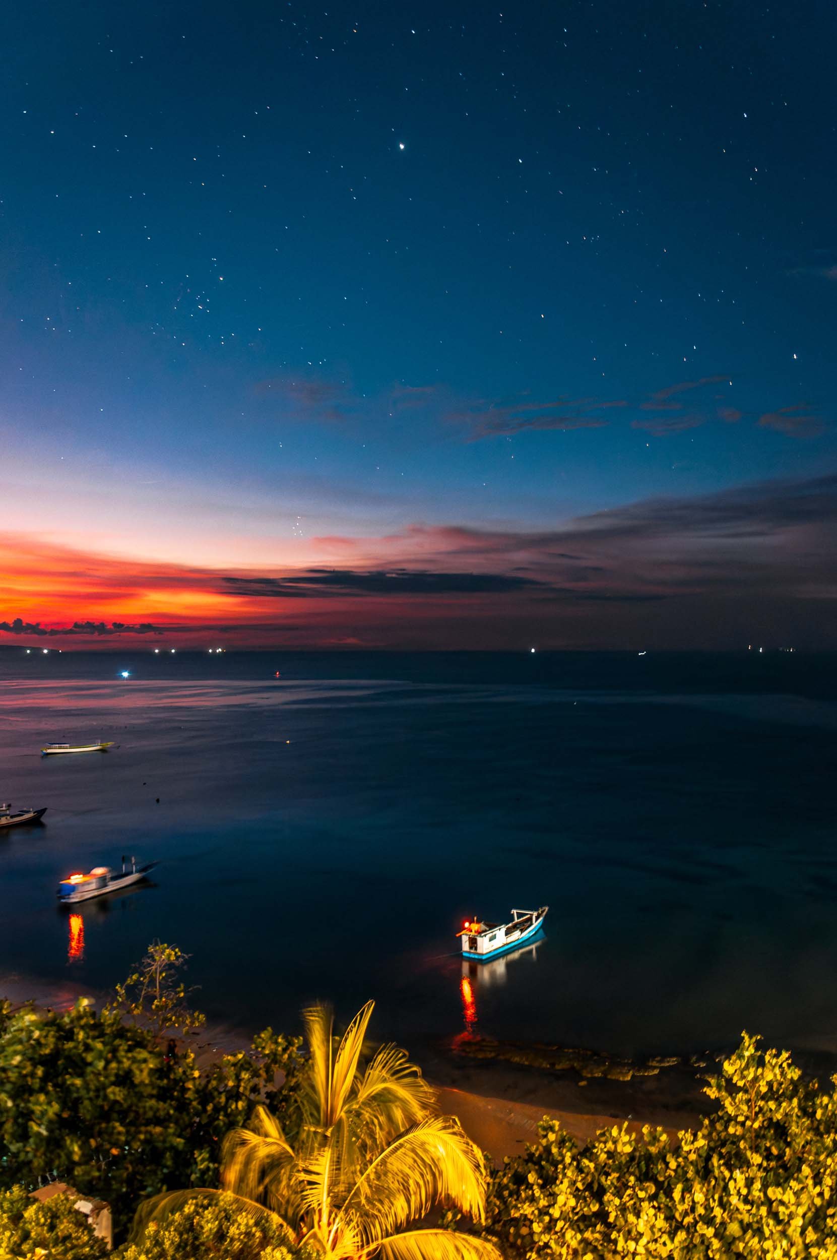 Nighttime scene of a calm ocean with several boats anchored, colorful sunset on the horizon, and stars appearing in the clear sky, with foliage in the foreground.