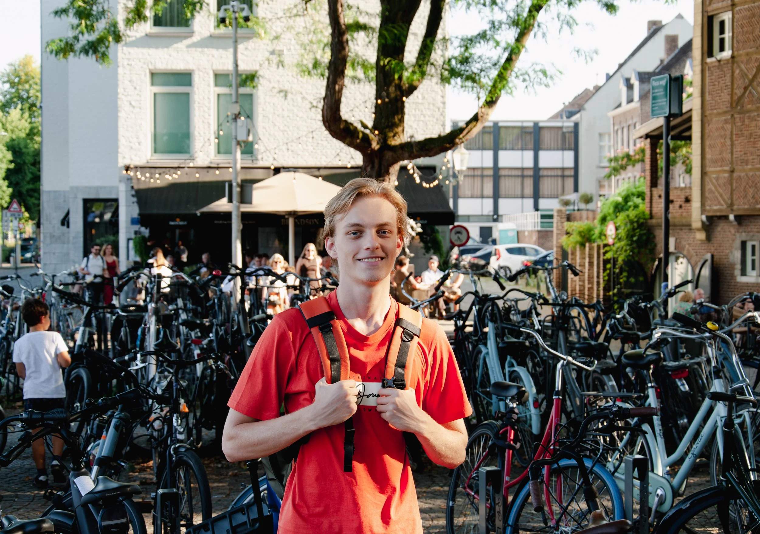 A young man in a red t-shirt with a backpack standing in front of a bike rack filled with bicycles, in an outdoor urban setting with buildings, trees, and people in the background.