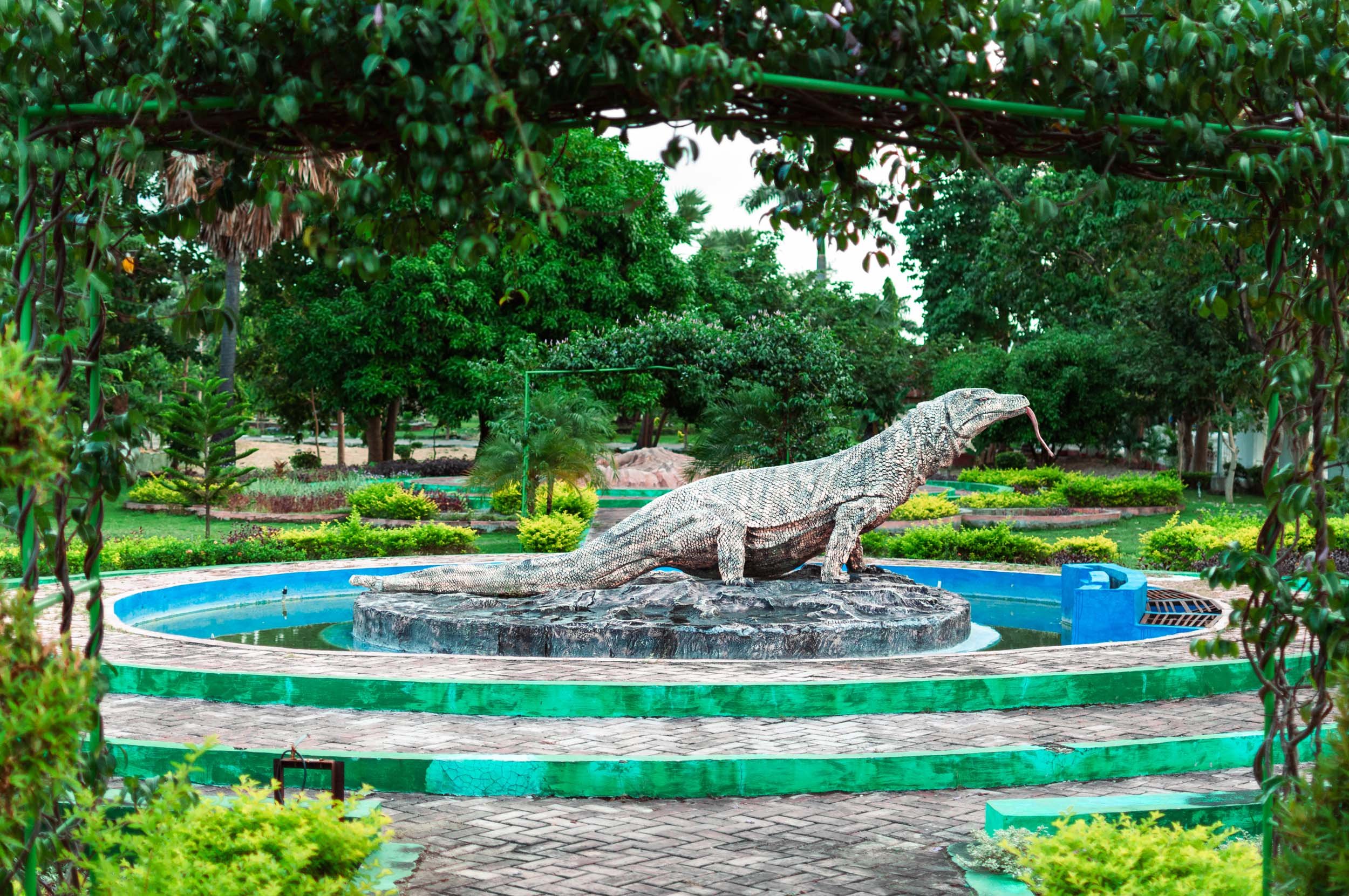 Statue of a large iguana in a circular fountain surrounded by lush greenery in a park.