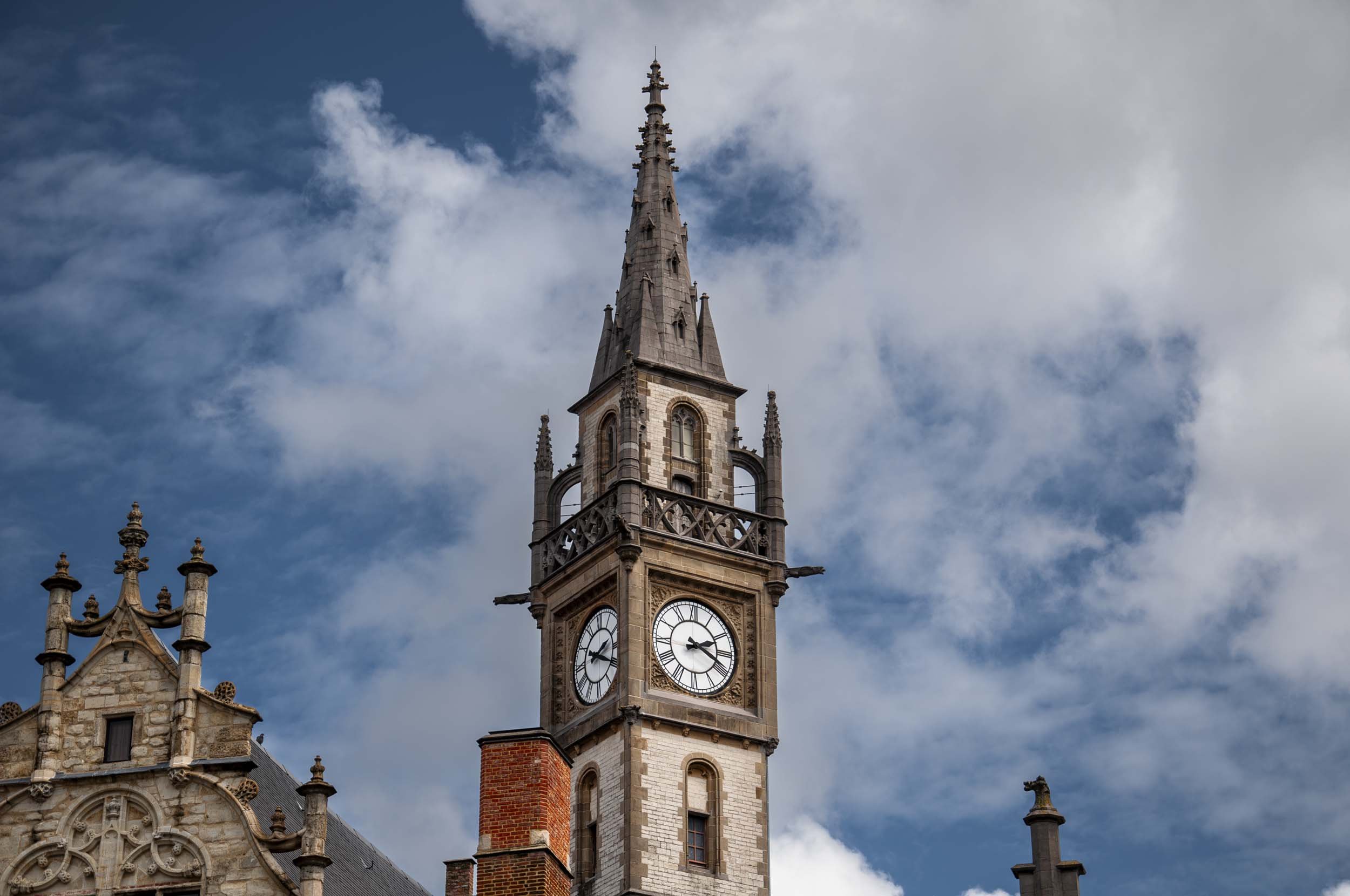 A historic clock tower in Ghent, with a gothic spire against a cloudy sky, part of an old stone building with ornate architectural details.