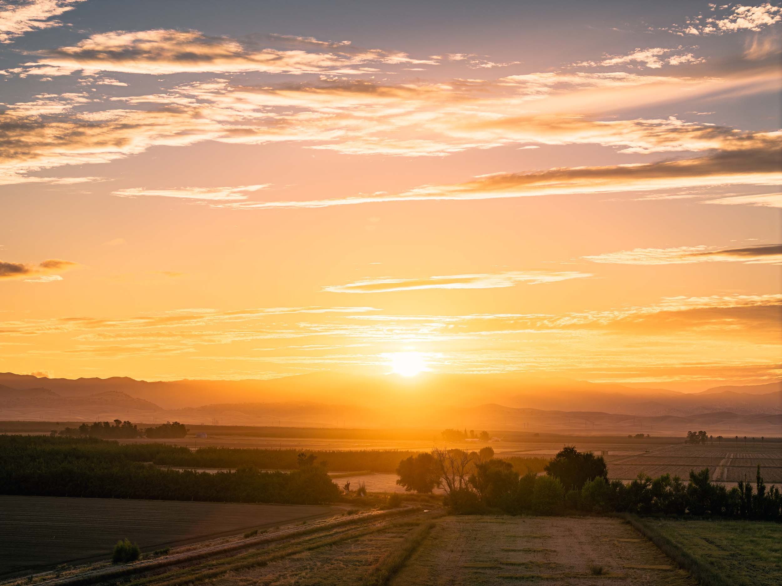 Sunset over a rural landscape with open fields, trees, and mountains in the distance, warm orange and yellow hues in the sky with scattered clouds.