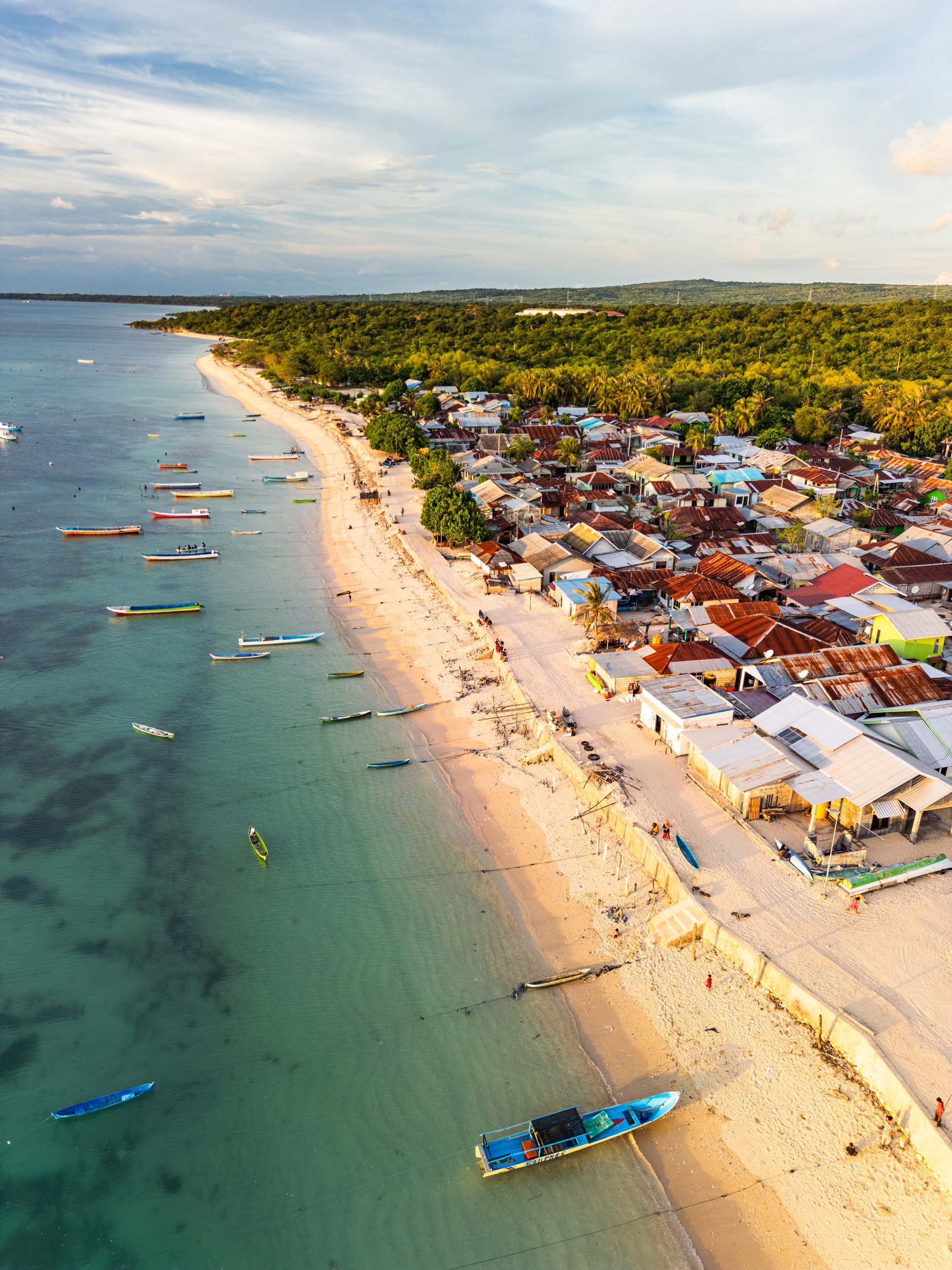 Aerial view of a coastal village with colorful houses, boats docked along the sandy beach, and lush green hills in the background under a partly cloudy sky.