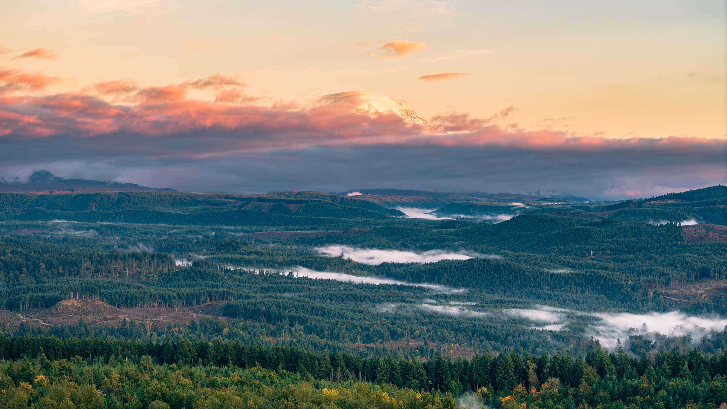A scenic view of a mountain landscape during sunrise or sunset, featuring rolling hills covered in dense green forests, with some mist or low clouds lingering in the valleys, and a sky with colorful clouds illuminated by the sun.