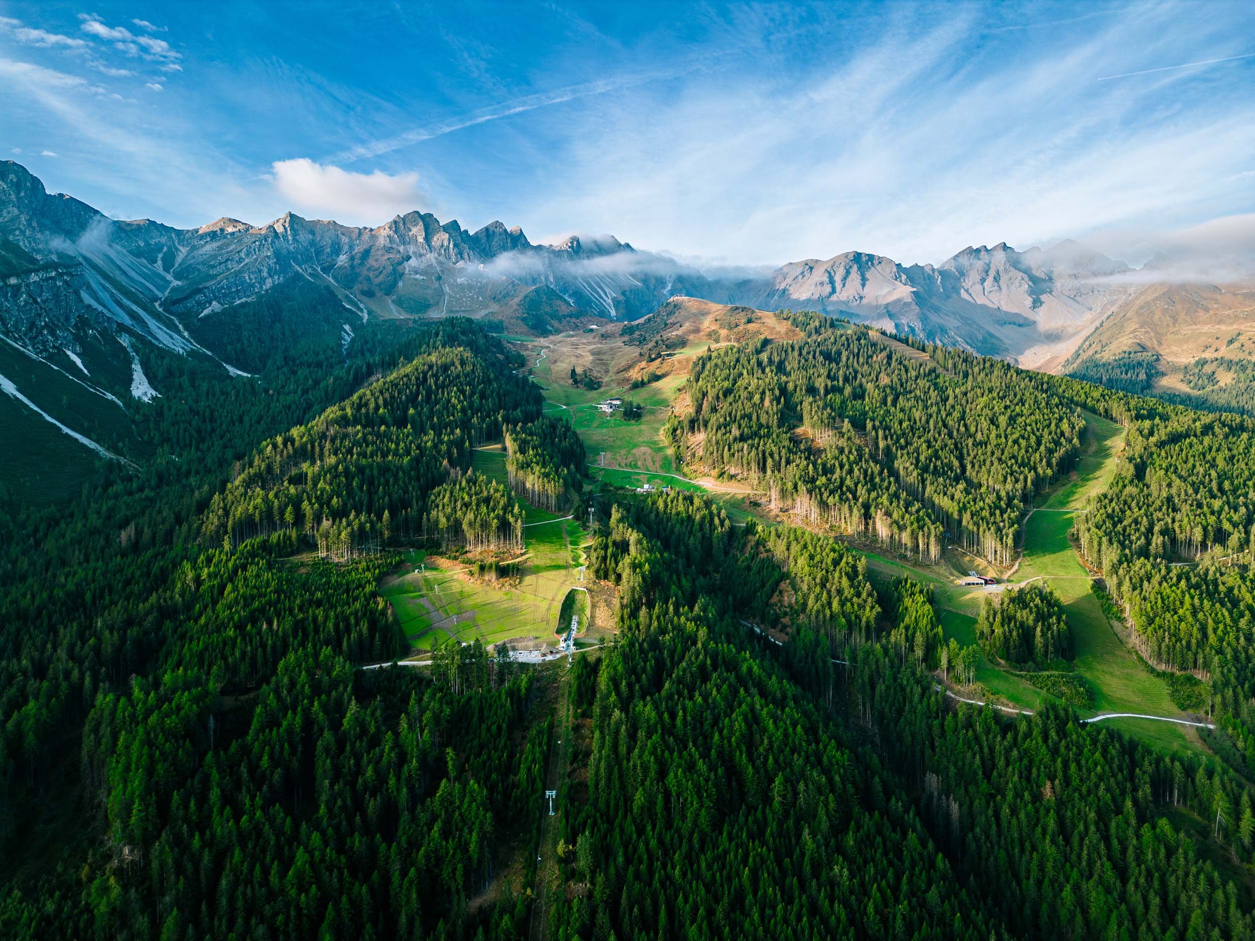 Aerial view of landscape in Alps with green pine forests, grassy clearings, and a small building, with rugged mountain peaks in the background and a blue sky with wispy clouds.