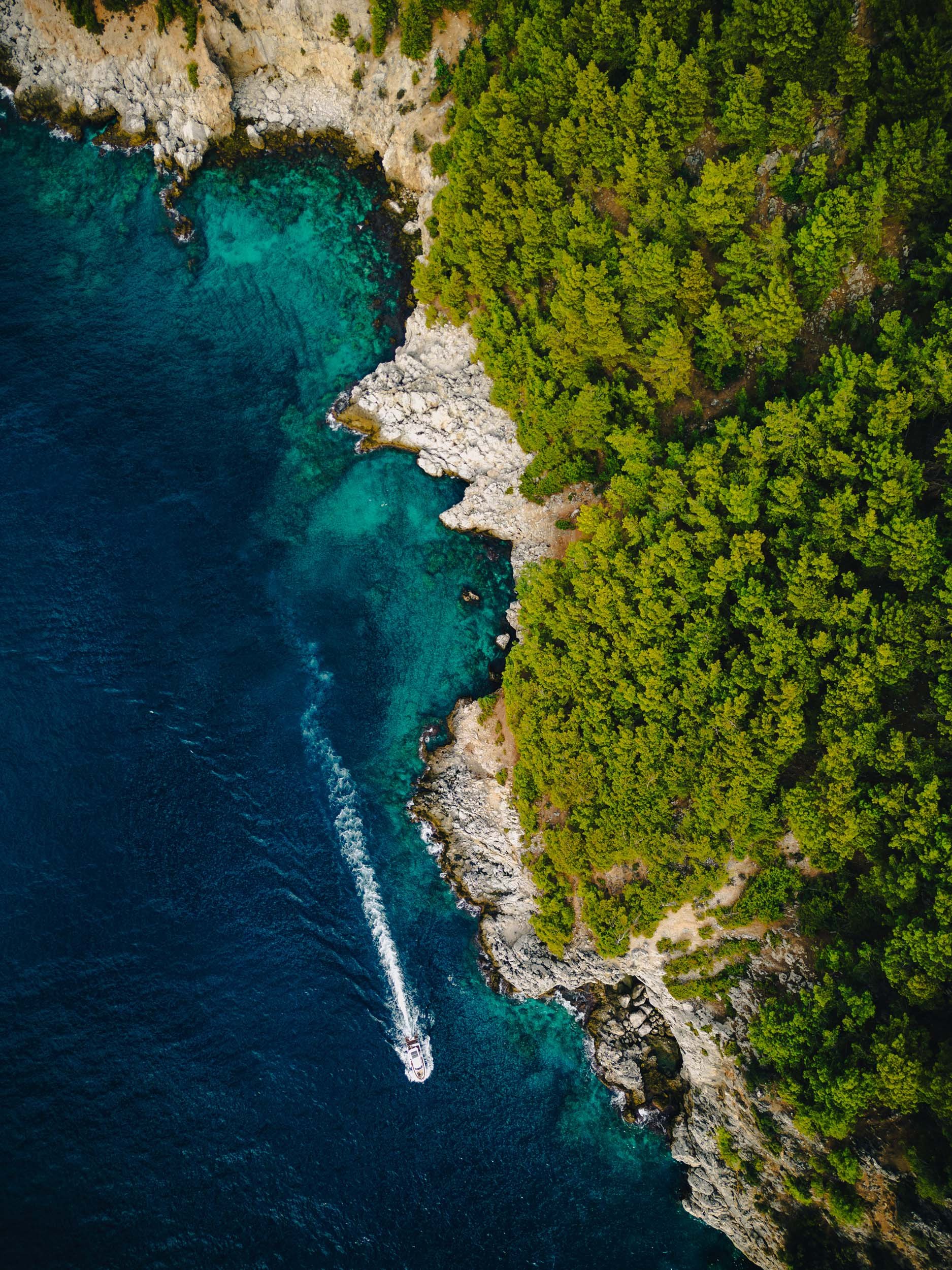Alanya aerial view of a boat sailing along a rugged shoreline with clear turquoise water, rocky cliffs, and dense green trees.