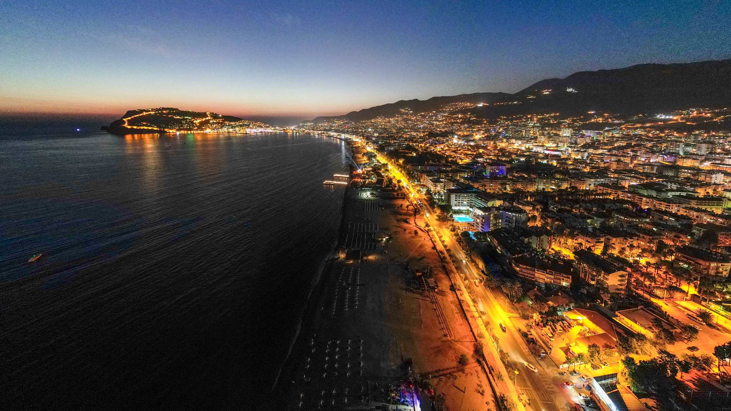 Night aerial view of a Alanya by the ocean with illuminated streets, buildings, and a beachfront, with hills in the background.