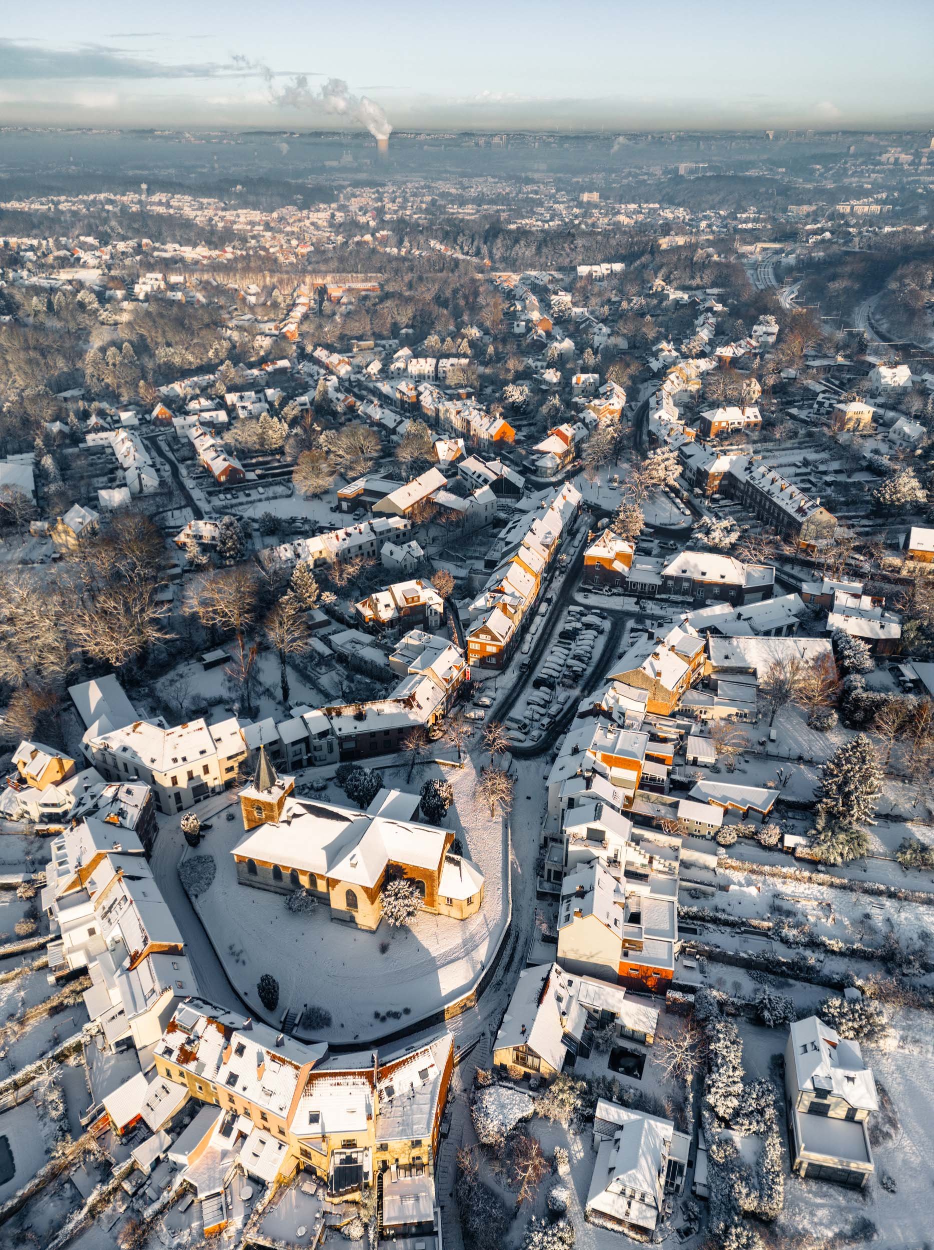 Linkebeek aerial view of a snow-covered town with a church in the center, surrounded by residential houses, trees, and winding roads, with a smoke plume rising in the distance.