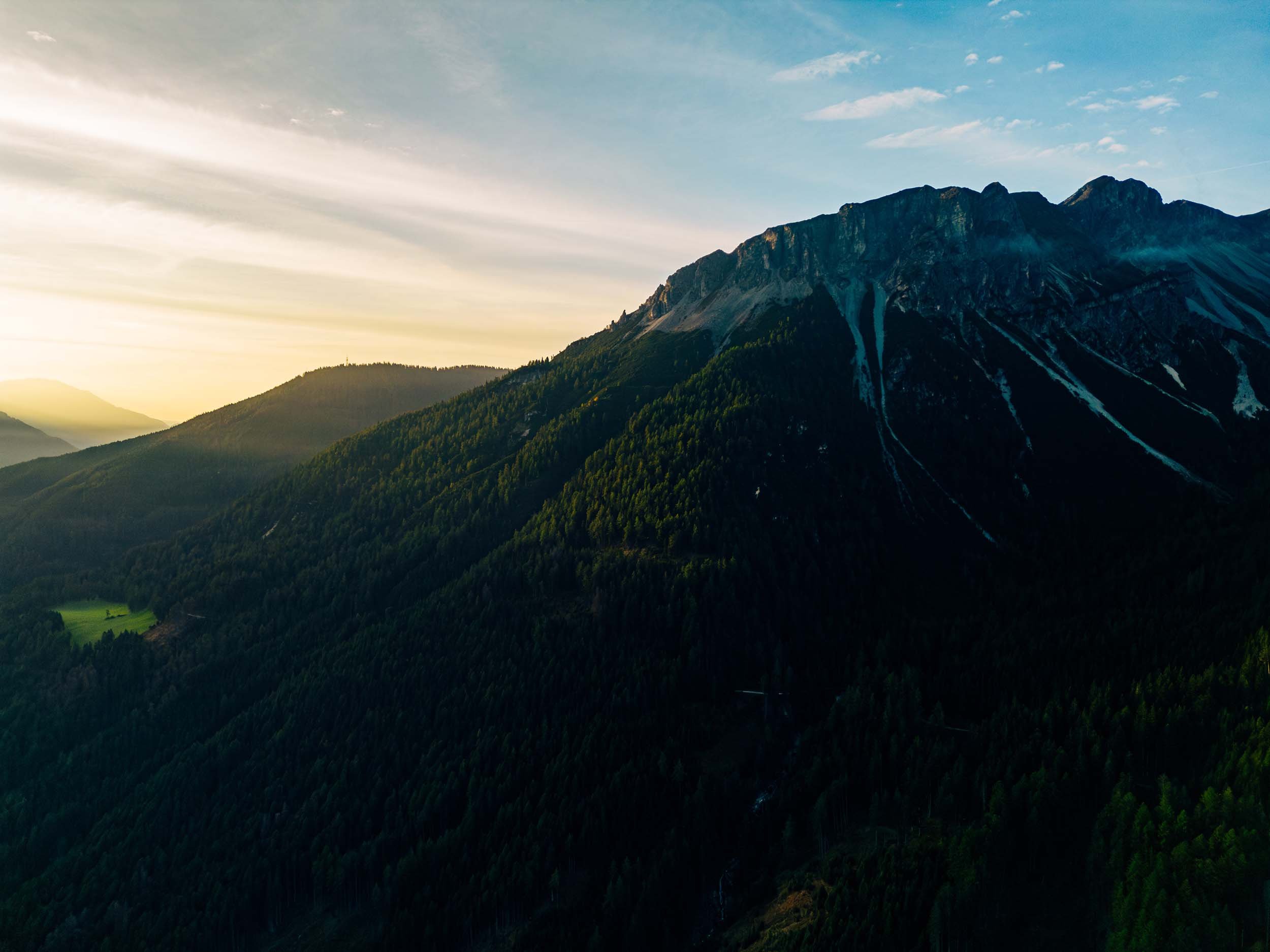 Sunset over Alps, covered with green forests, with a rugged peak in the background.