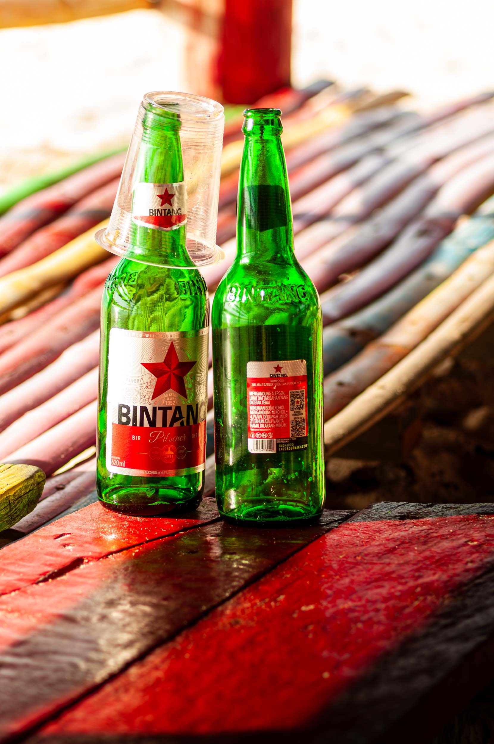 Two green glass bottles of Bintang beer, one with a clear plastic cup on top, placed on a rustic wooden surface outdoors.