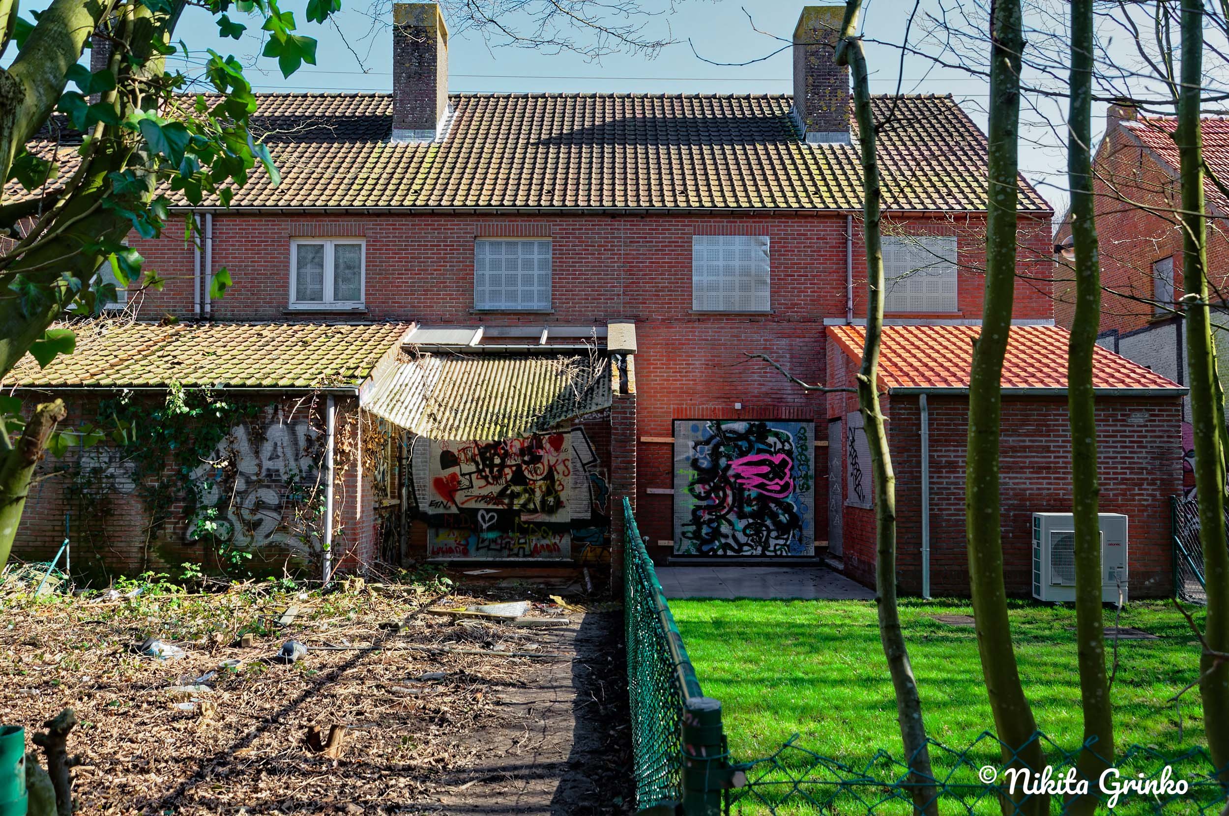 Backyard of a house with a garden, graffiti-covered shed, brick house with windows, a small lawn, trees, and an air conditioning unit.