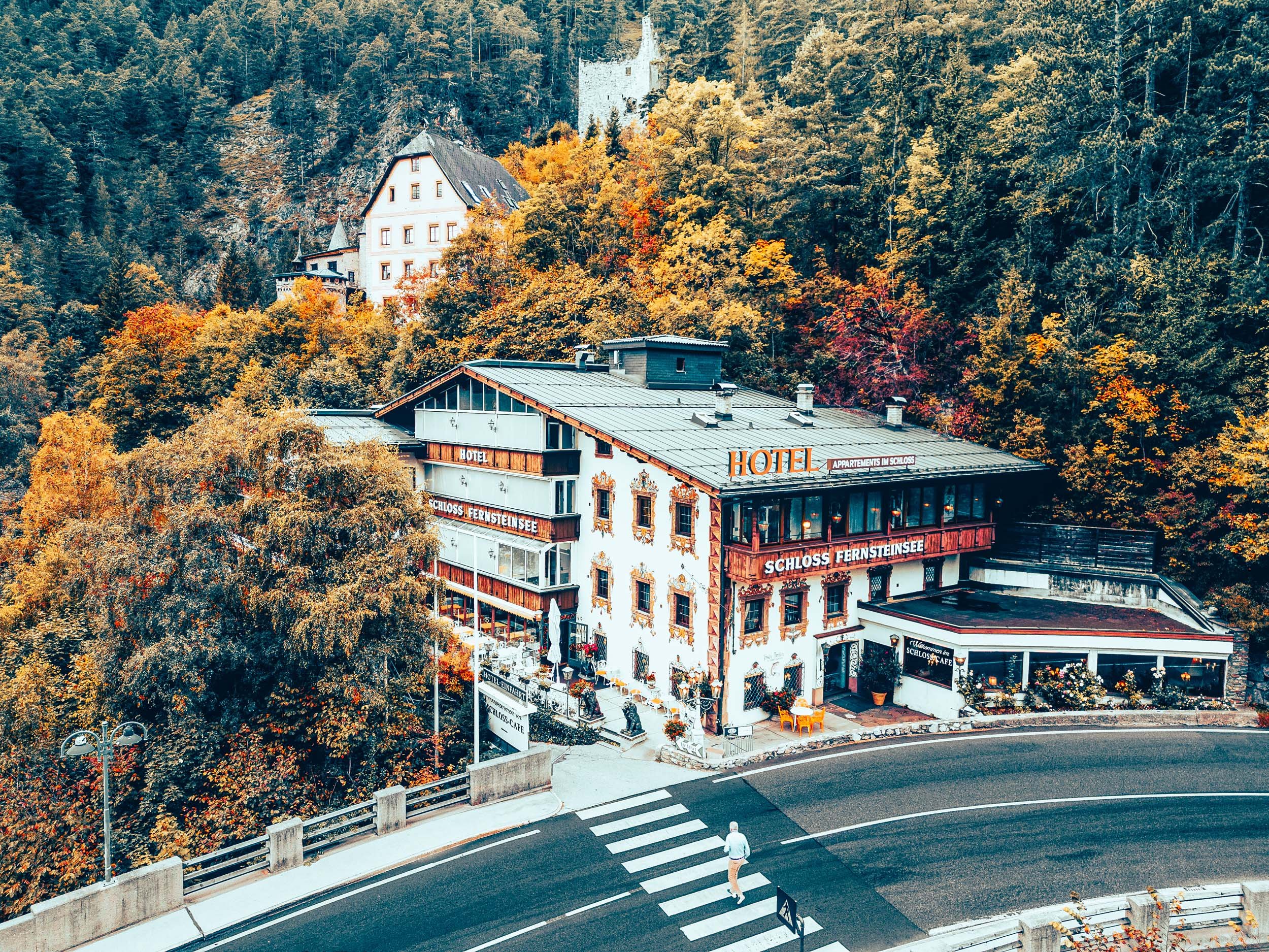 A hotel building on a hillside surrounded by colorful autumn trees, with a curved road and a person crossing a crosswalk in the foreground.