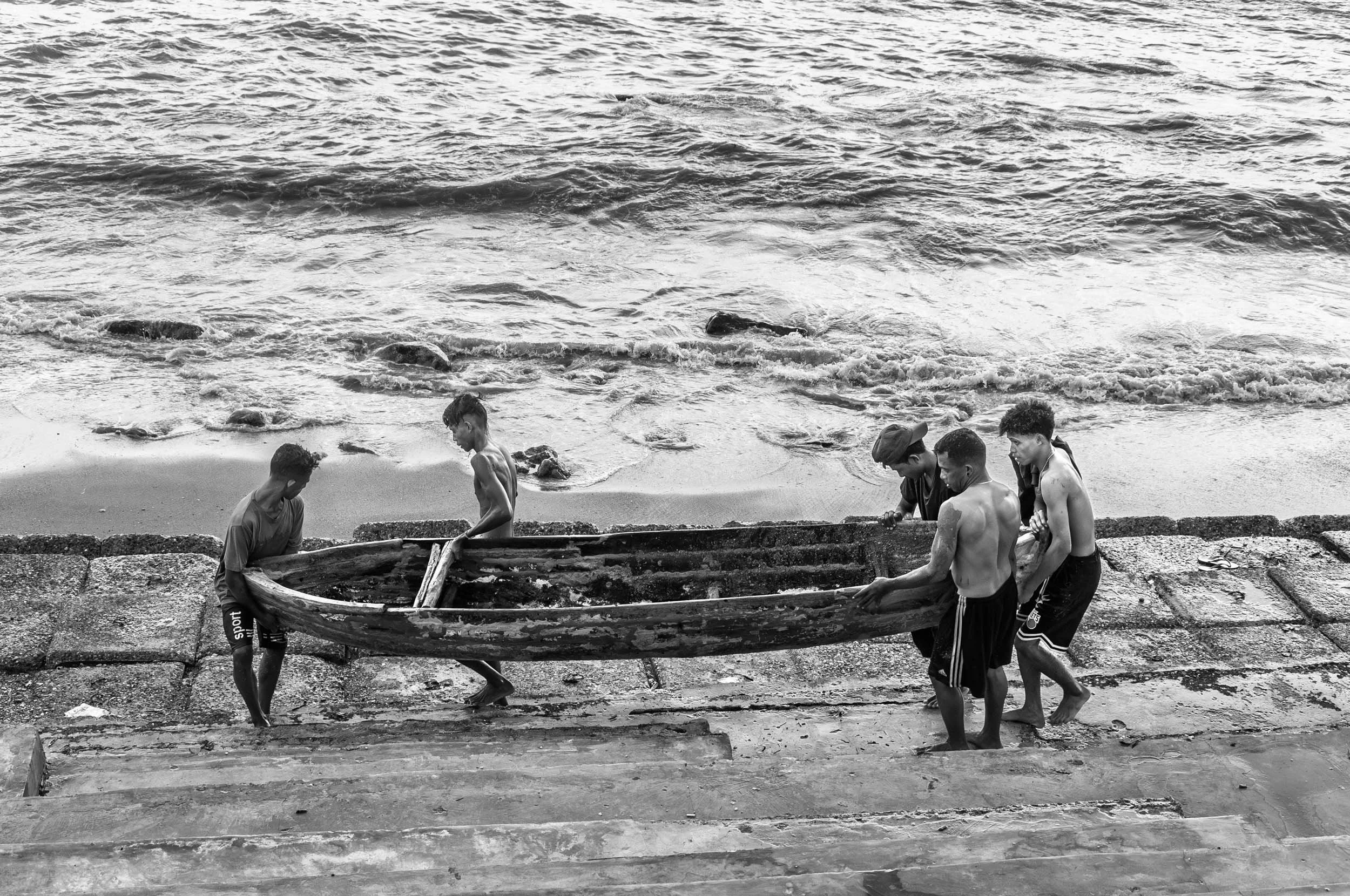 Five young boys carrying a wooden boat down a sandy and concrete staircase toward the ocean.
