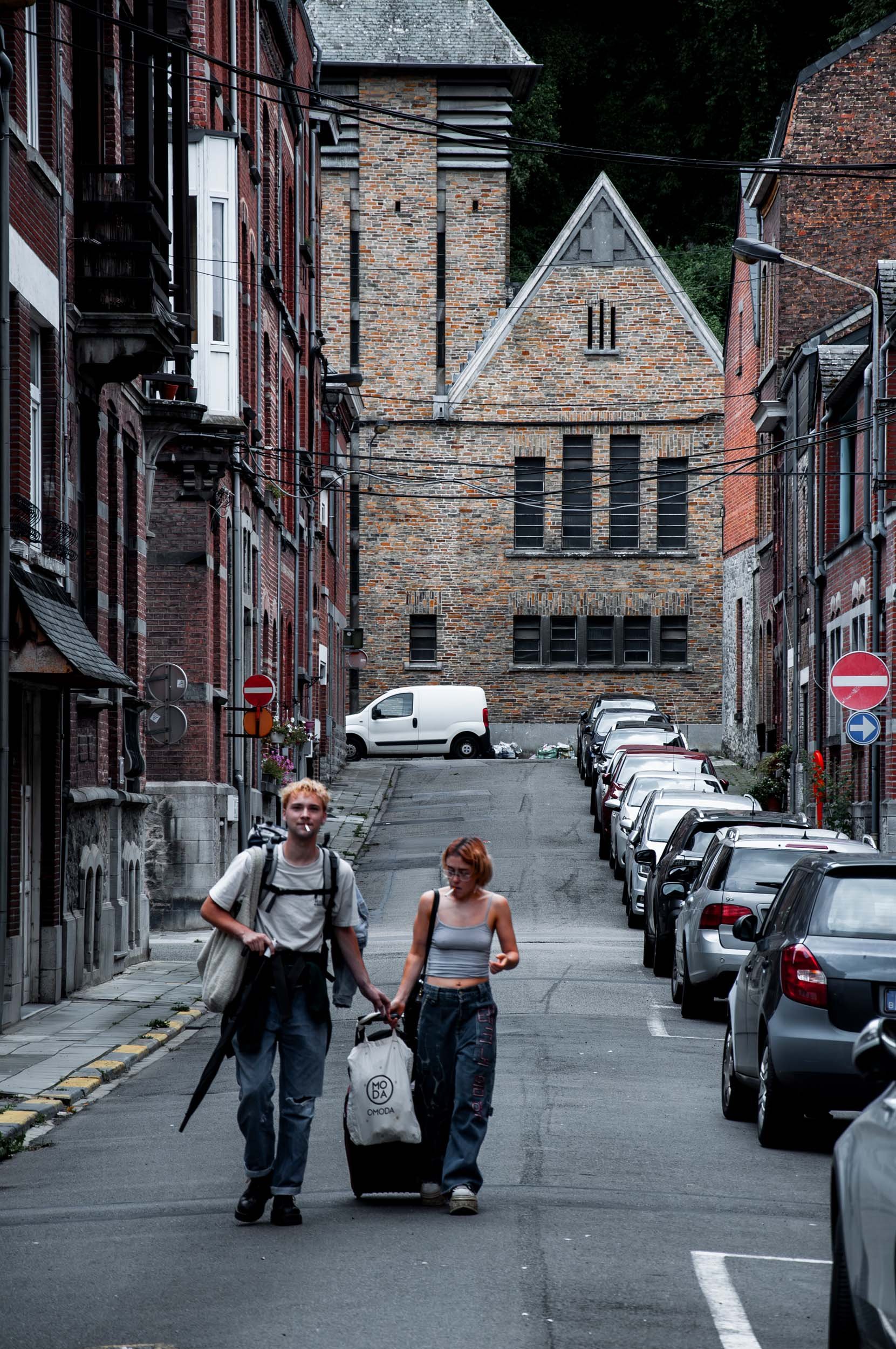 Two young people, a man and a woman, walk hand-in-hand down a city street with parked cars along the sides. The street is urban, lined with old brick buildings, and the background features a large brick building with gothic architectural elements. Th