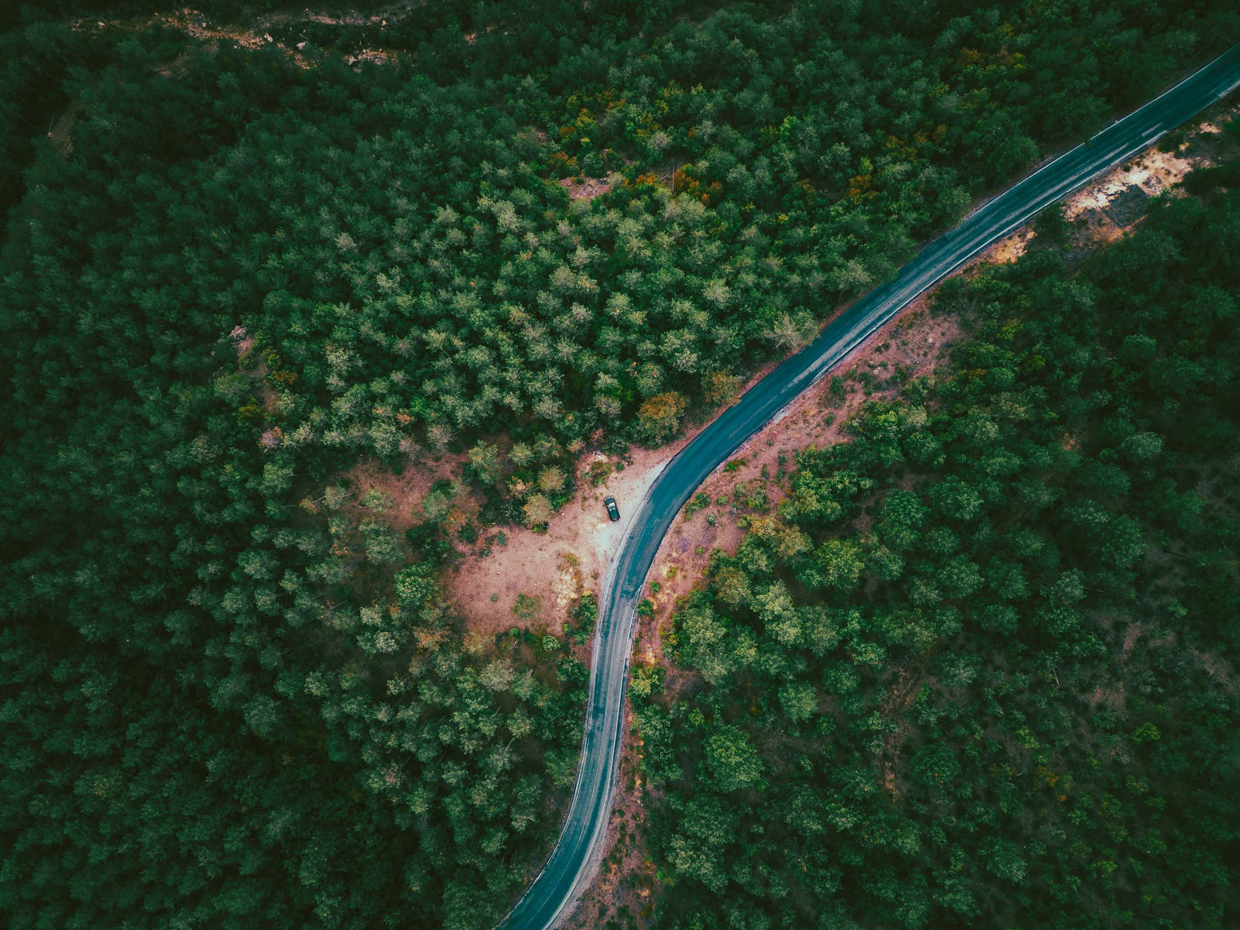 An aerial view of a winding road through dense green forest with a single parked vehicle on a small dirt patch beside the road.