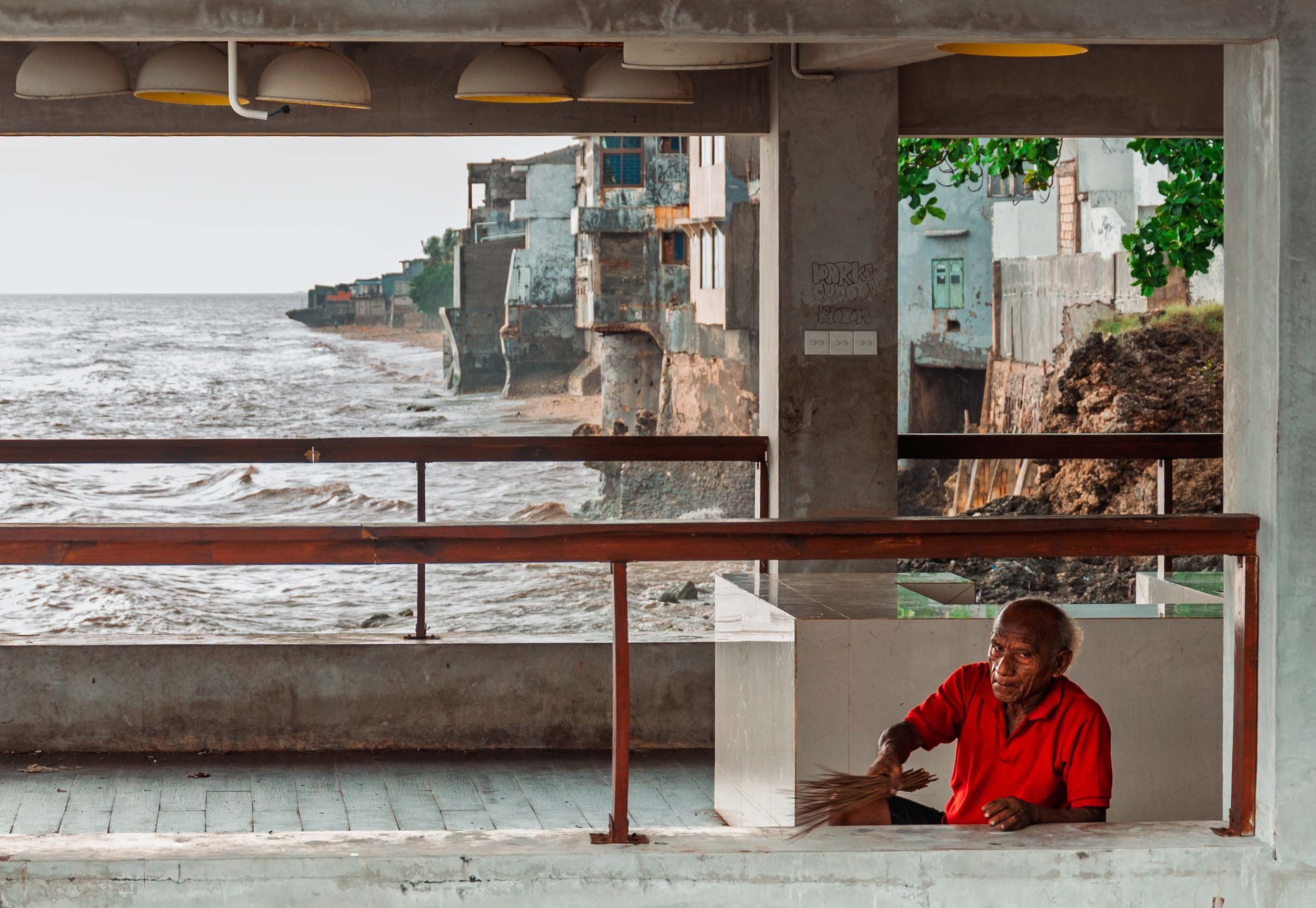 An elderly man in a red shirt sweeping on a balcony overlooking the ocean and old buildings on the coastline.