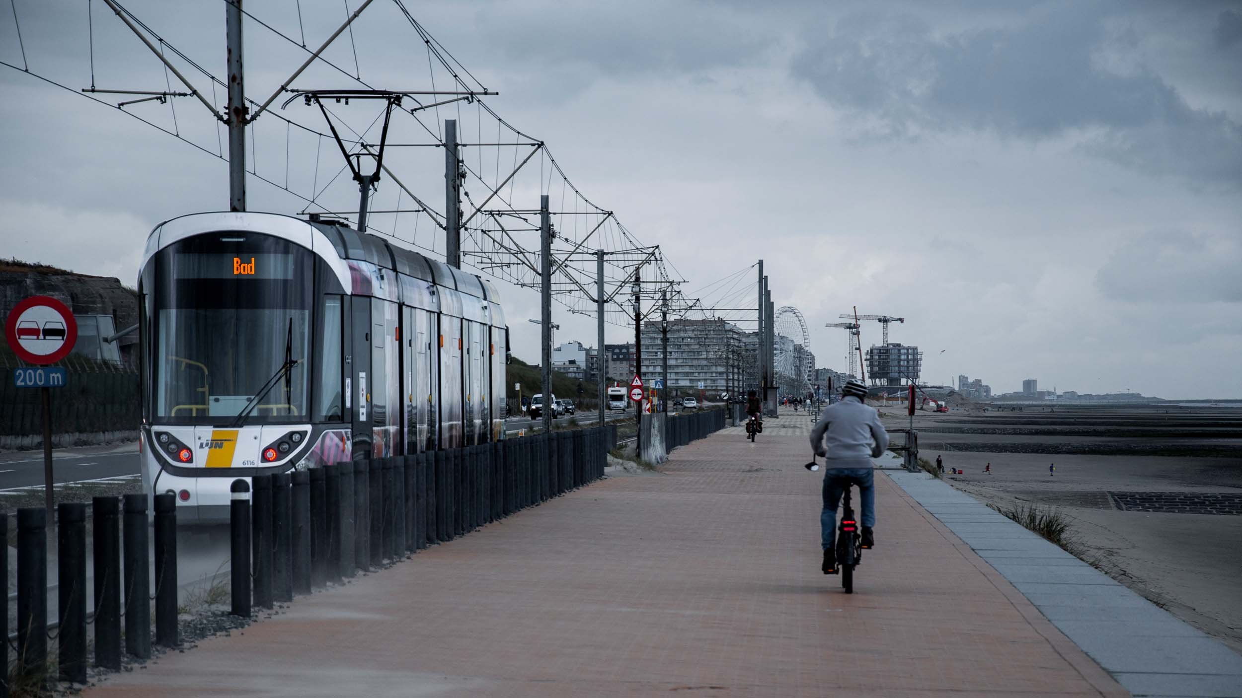 A coastal walkway with a cyclist riding along the paved path under cloudy skies. A modern tram is traveling nearby on tracks separated by a black fence, with overhead electrical wires. In the background, there are high-rise buildings and construction