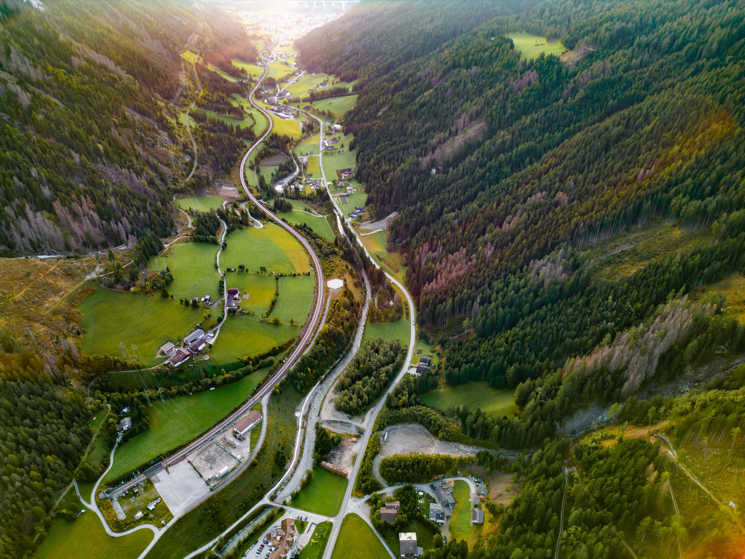 Aerial view of a winding road through a lush green valley with small houses, surrounded by forested mountains and fields.