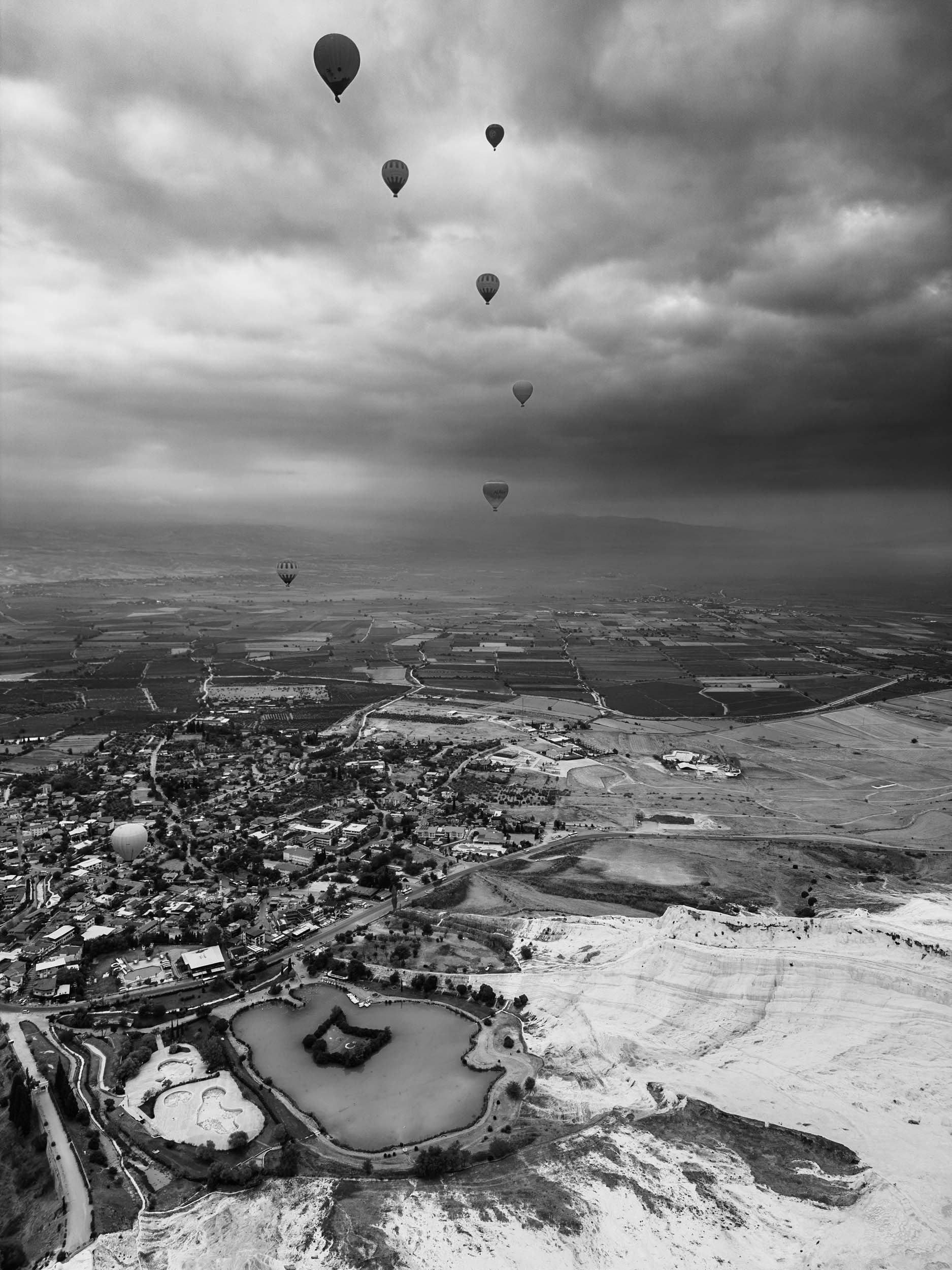 Seven hot air balloons floating over a landscape with a town, farmland, and a salt lake, under a cloudy sky.