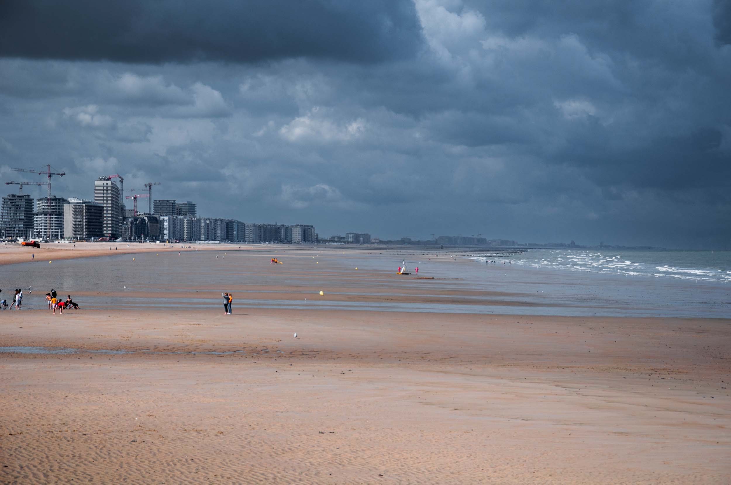 A beach under dark, cloudy skies with high-rise buildings in the background, some construction cranes visible, and people walking near the shoreline.