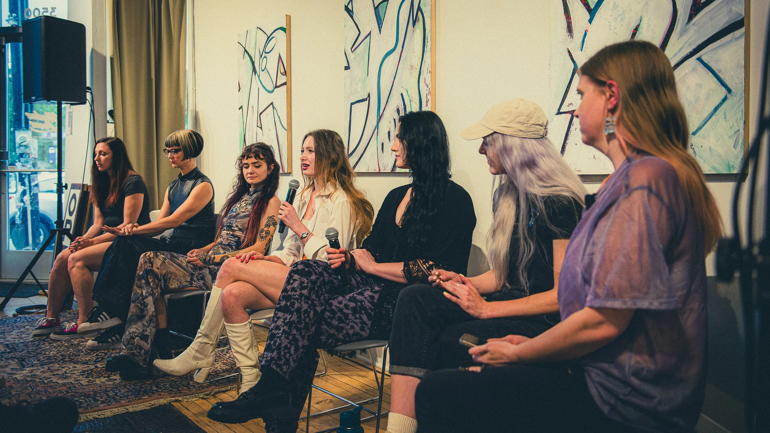 A group of women sitting on stage, some holding microphones, with abstract artwork on the wall behind them.
