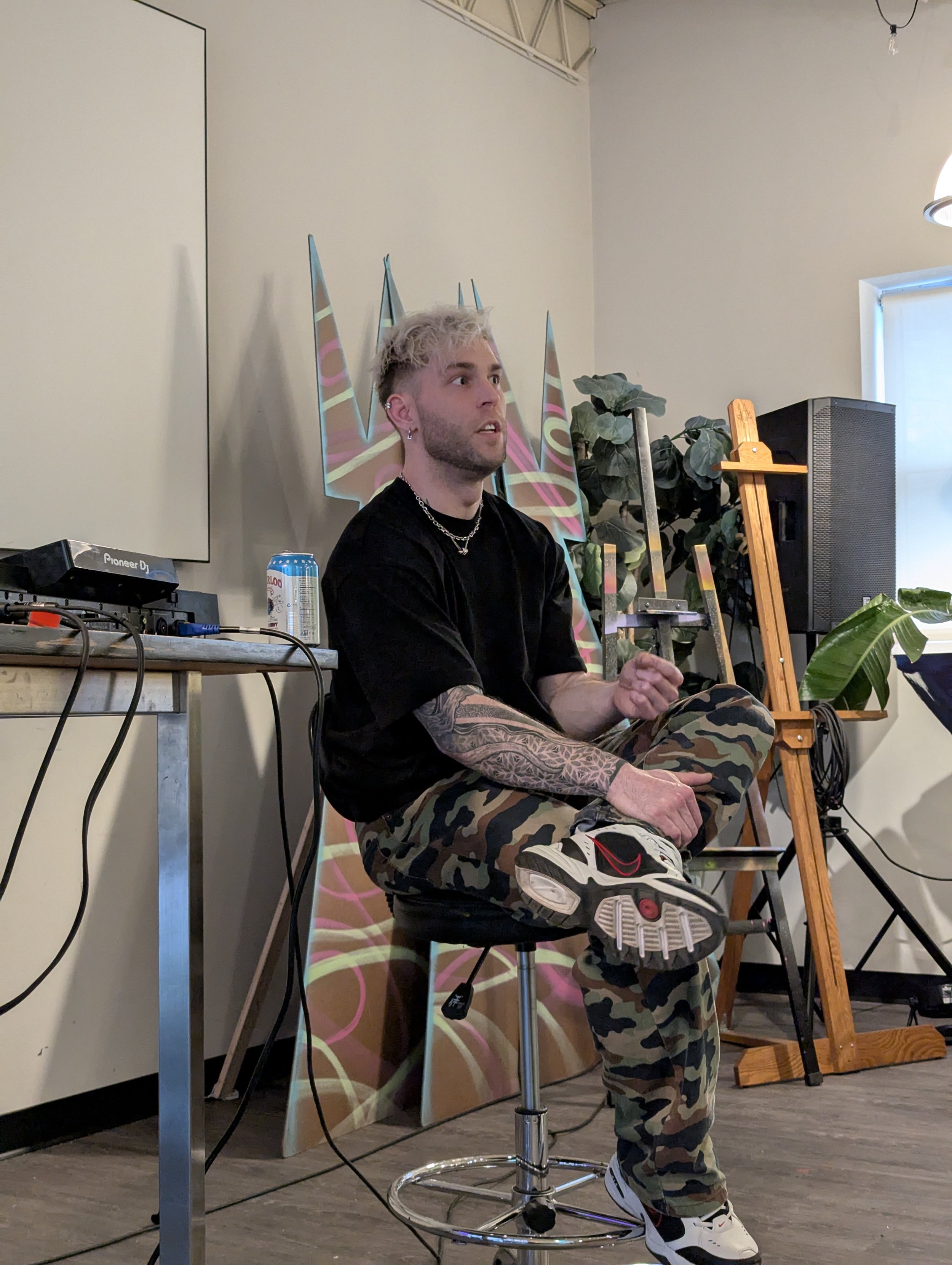A young man with bleached hair, tattoos, and earrings sitting on a stool, wearing camouflage pants and sneakers, in a room with art supplies and a DJ setup.