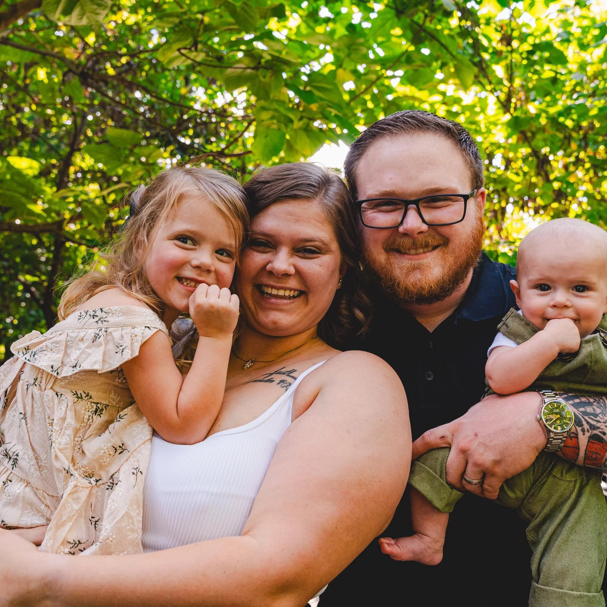 A happy family of four smiling and posing outdoors under green trees. The mother holds a young girl while the father holds a baby.