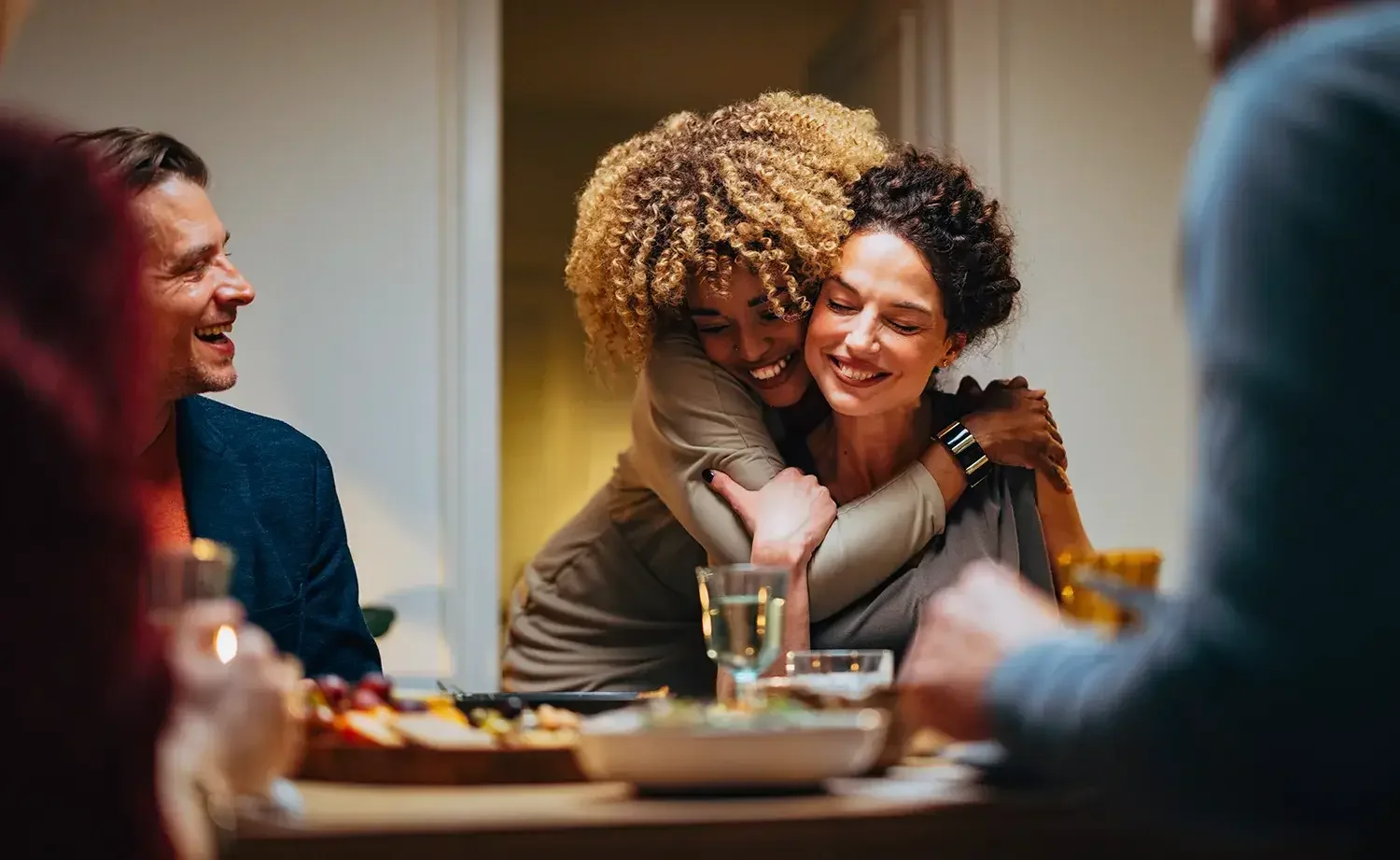 A candid photo of two smiling women hugging while seated at a dining table with friends and food.