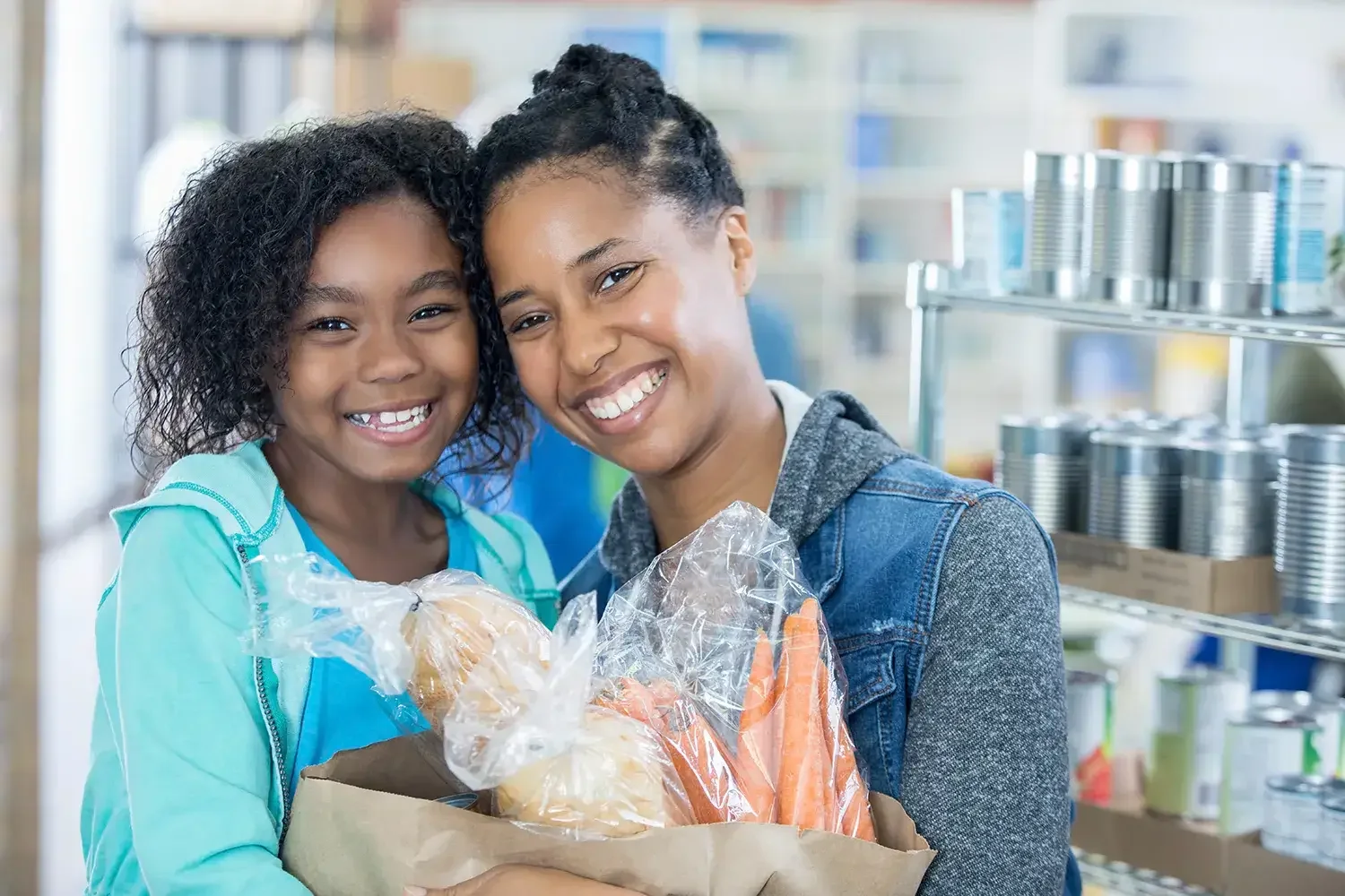 A smiling Black woman and young girl embrace while holding a grocery bag filled with carrots and other food items inside a food pantry.