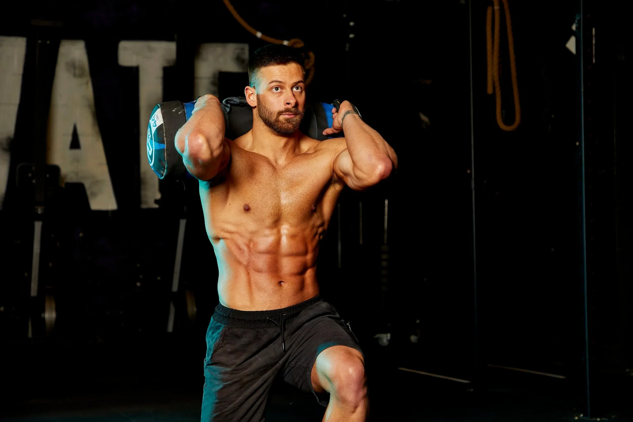A muscular man with a beard, shirtless, holding a weighted sandbag on his shoulders, performing a workout in a gym with black walls and exercise equipment in the background.