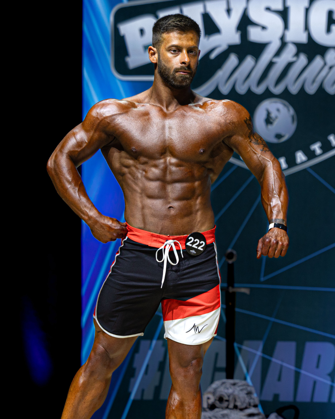 A muscular man with tanned skin, dark hair, and a beard, posing on stage at a fitness competition. He is wearing black and red shorts with a number 222 tag on his waist and a black wristband, with his hands on his hips. There is a backdrop with fitness-related text and graphics.