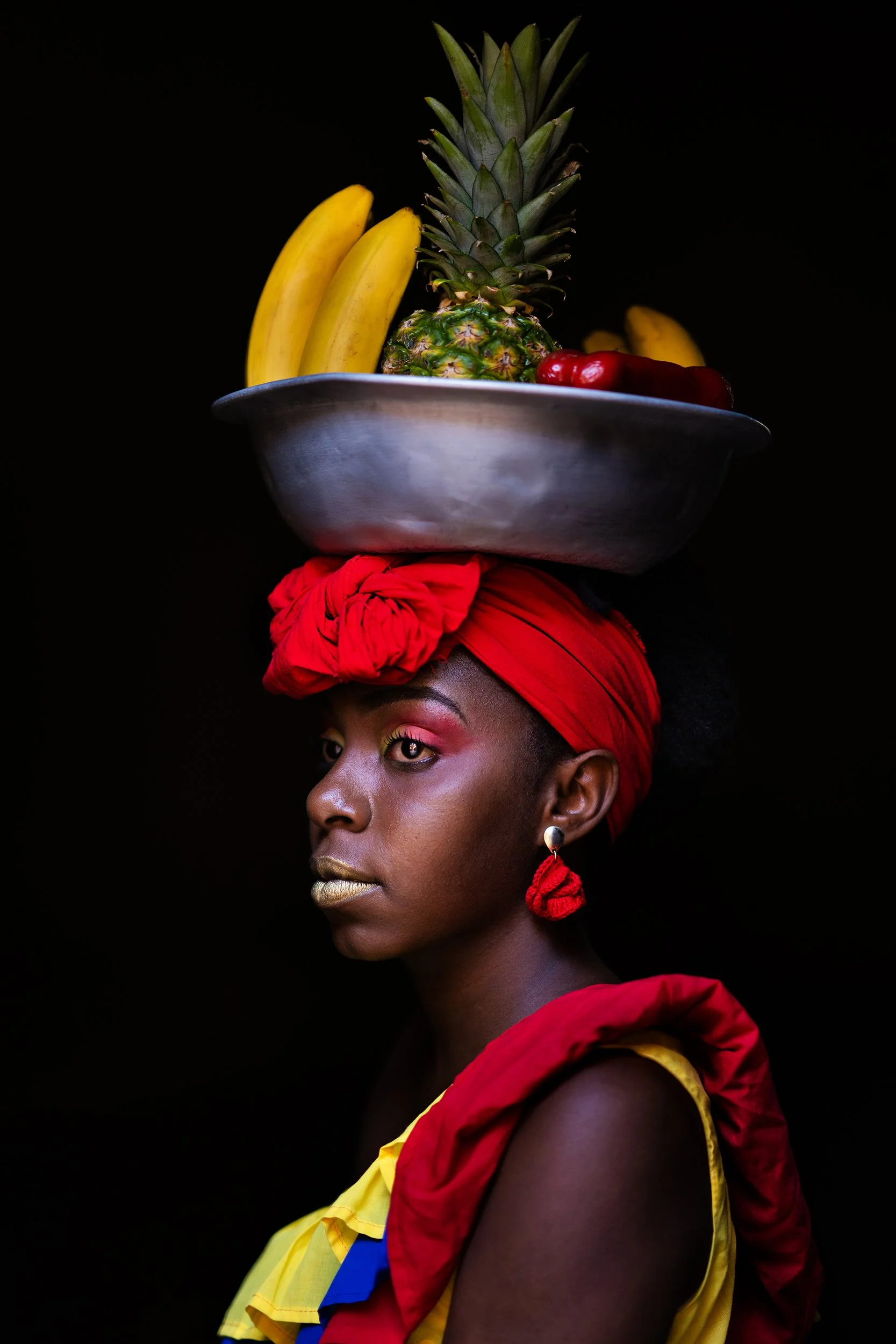 A woman with dark skin wearing a red headwrap, yellow and red clothing, and gold makeup, balances a tray of tropical fruits including a pineapple and bananas on her head against a black background.