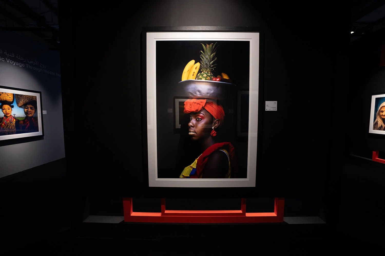 Photograph of a dark-themed gallery wall featuring a portrait of a woman with dark skin, wearing a headwrap and earrings, with a bowl of fruit balanced on her head, including a pineapple, bananas, and berries.