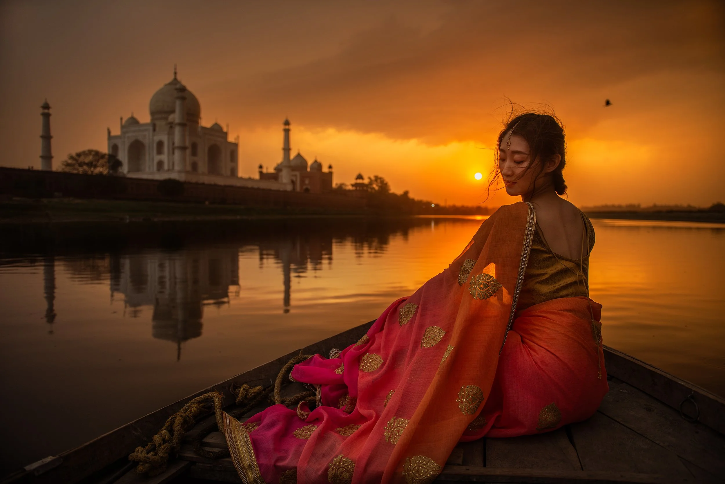 A woman in traditional Indian attire sitting in a boat on a river during sunset, with the Taj Mahal in the background.