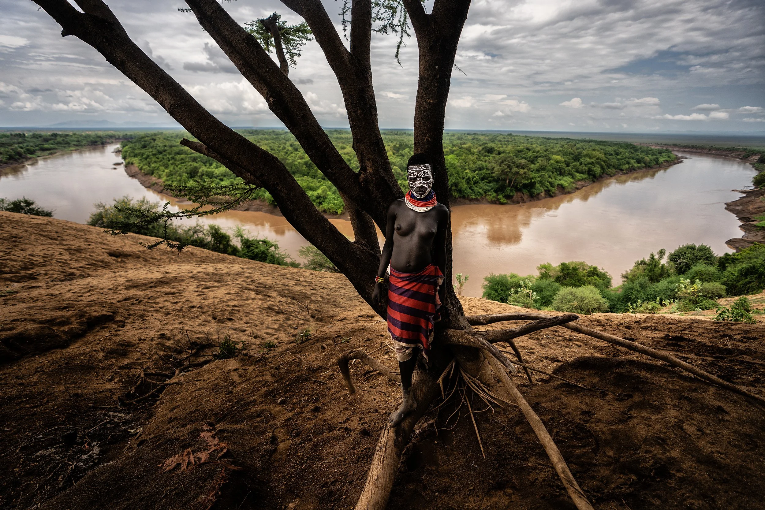 A woman in traditional Maasai attire and face paint standing by a tree overlooking a river in a savannah landscape under cloudy skies in Africa.