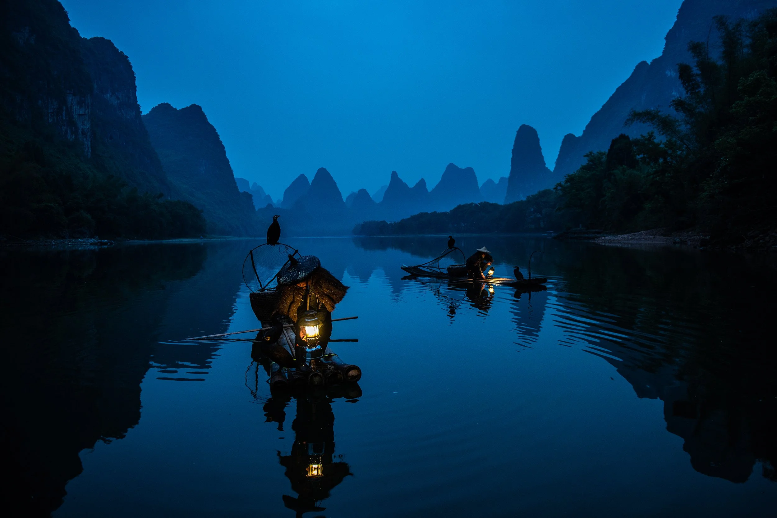 Three boaters on traditional bamboo boats with lanterns, floating on a serene river at dusk, surrounded by towering karst mountains covered in greenery.