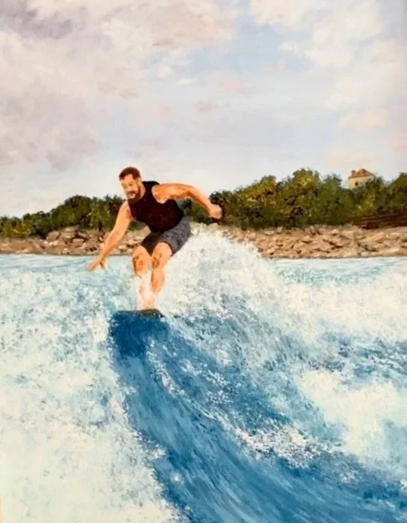 A man wake surfing on a blue water wave with a shoreline and trees in the background.