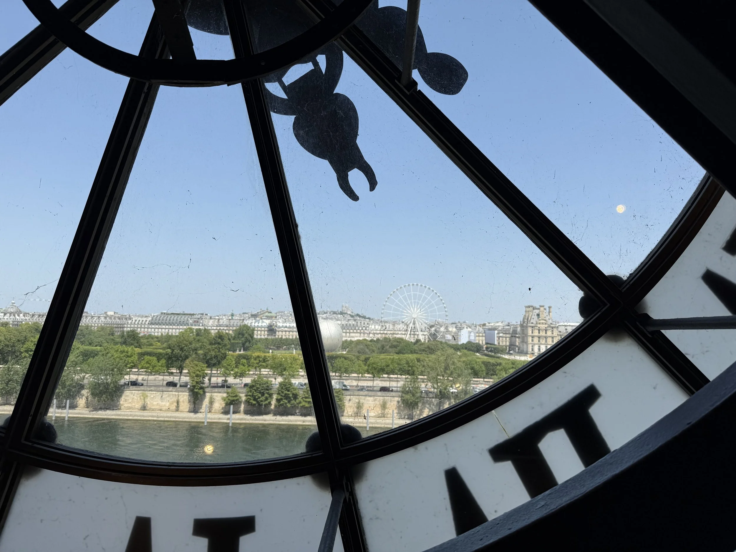View of Paris skyline through the clock face of the Musée d'Orsay, including the Seine River and a ferris wheel in the distance, with the moon visible in the sky.
