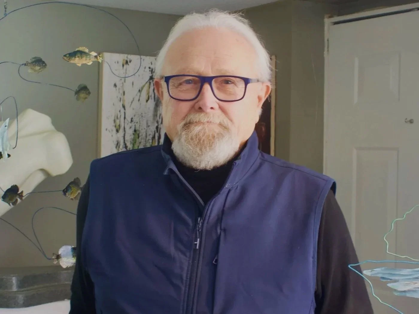 Older man with gray hair, beard, and glasses standing indoors, wearing a navy vest over a black shirt, with background art and a door.