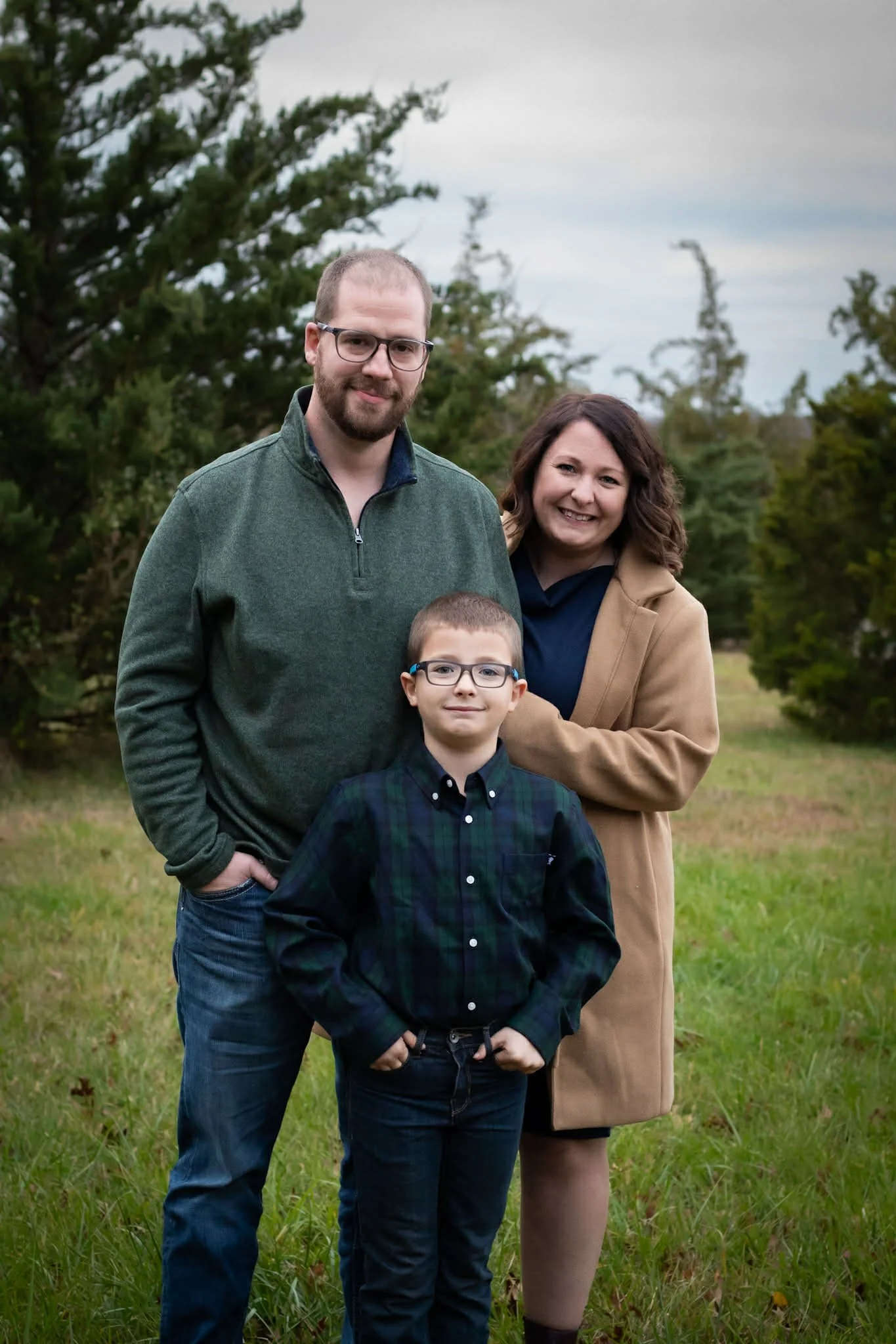 A family of three standing outdoors on a grassy field with trees in the background. The man has light skin, glasses, facial hair, and wears a green quarter-zip sweater. The woman has light skin, shoulder-length curly hair, and wears a tan coat. The boy has light skin, glasses, short hair, and wears a dark plaid button-up shirt.