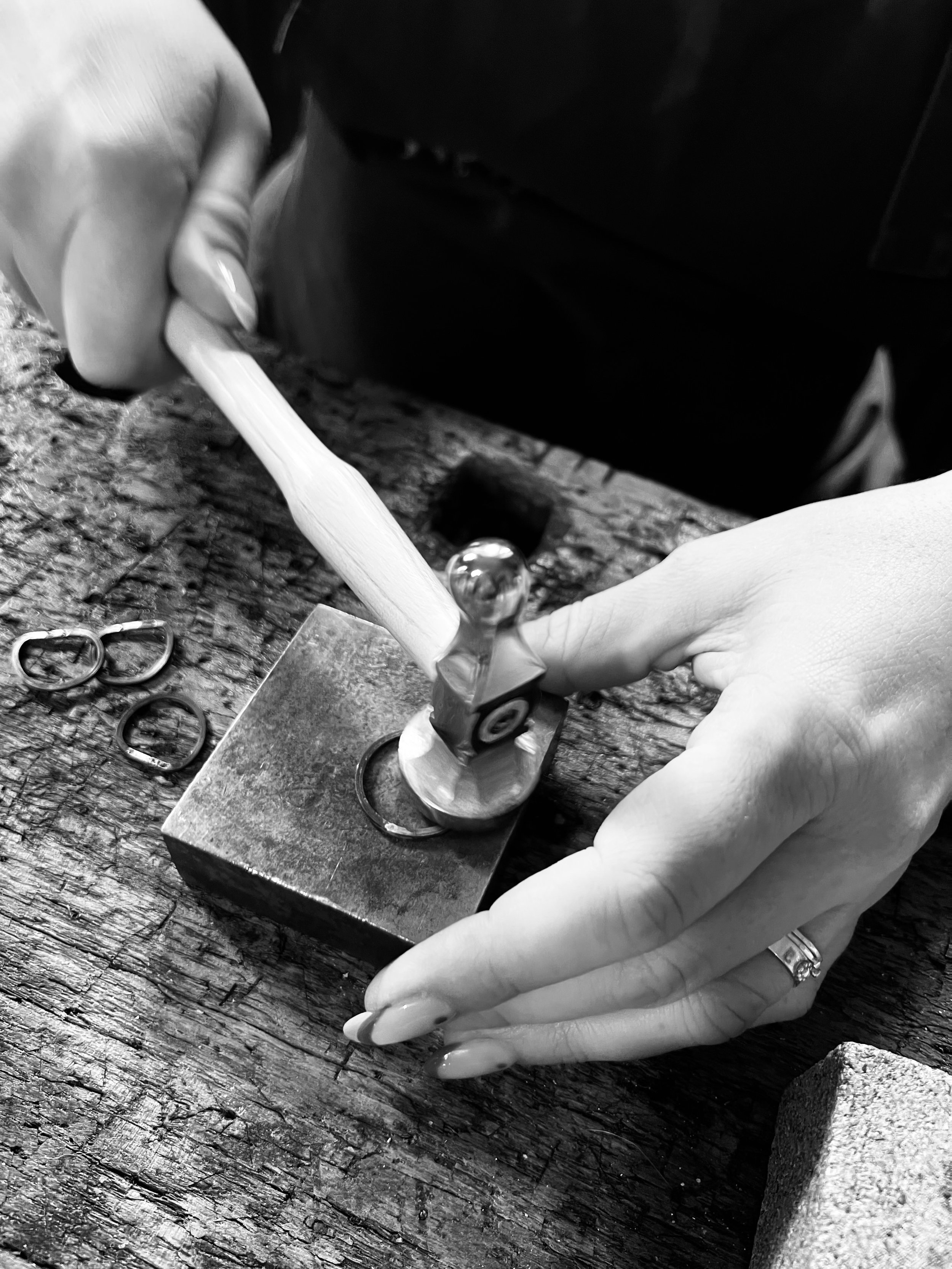 Close-up black and white photo of a person holding a small hammer with both hands, with the focus on their fingers and the hammer, in a workshop with tools and a wooden work surface.