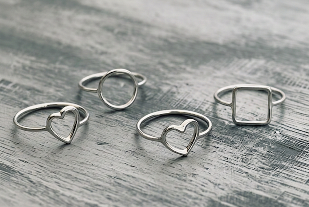 Five silver rings with various heart and square shapes, placed on a weathered wooden surface.