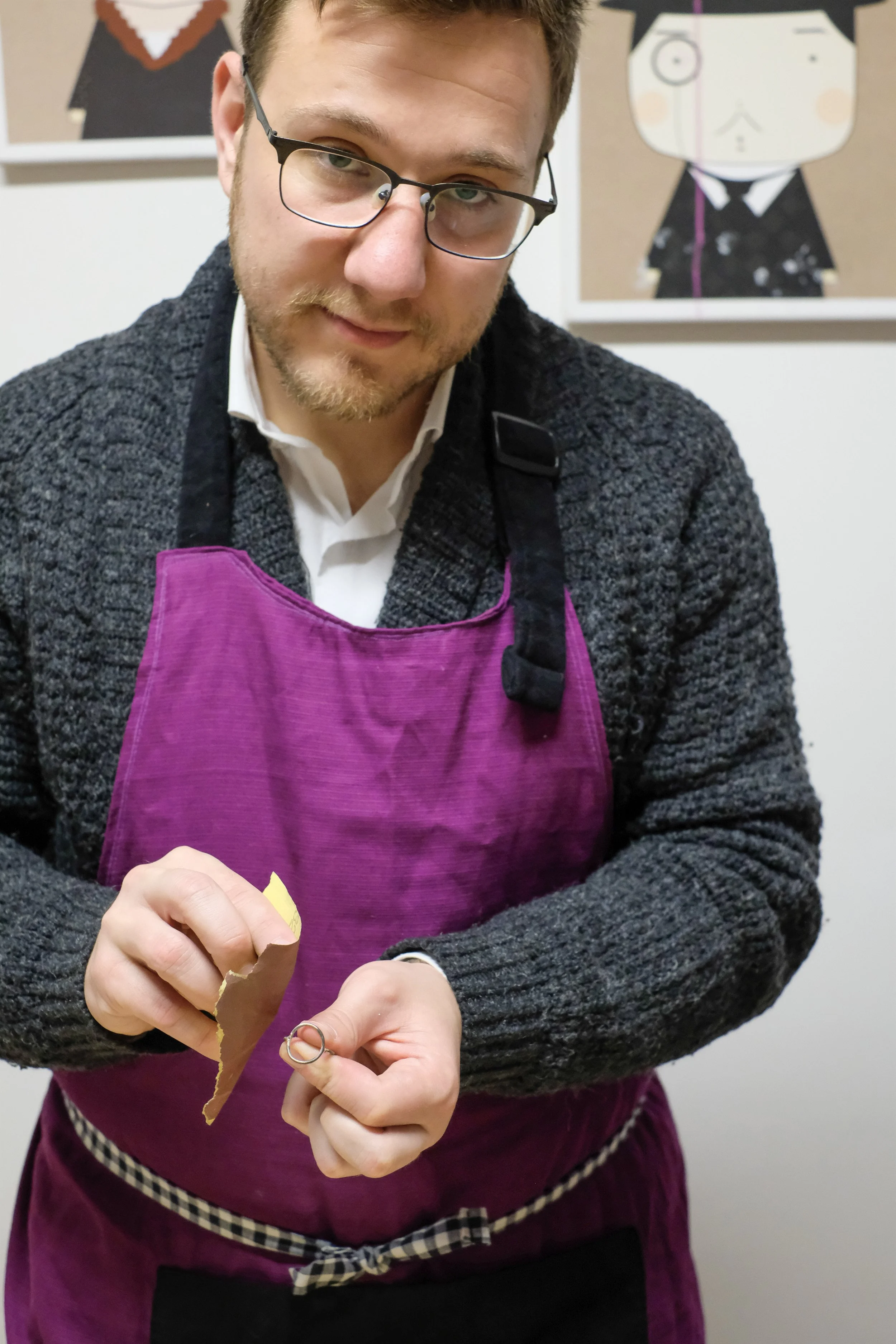 A man with glasses and a beard wearing a purple apron over a gray sweater, holding a piece of ripped brown paper or cardboard. The background has framed artwork depicting cartoon characters.