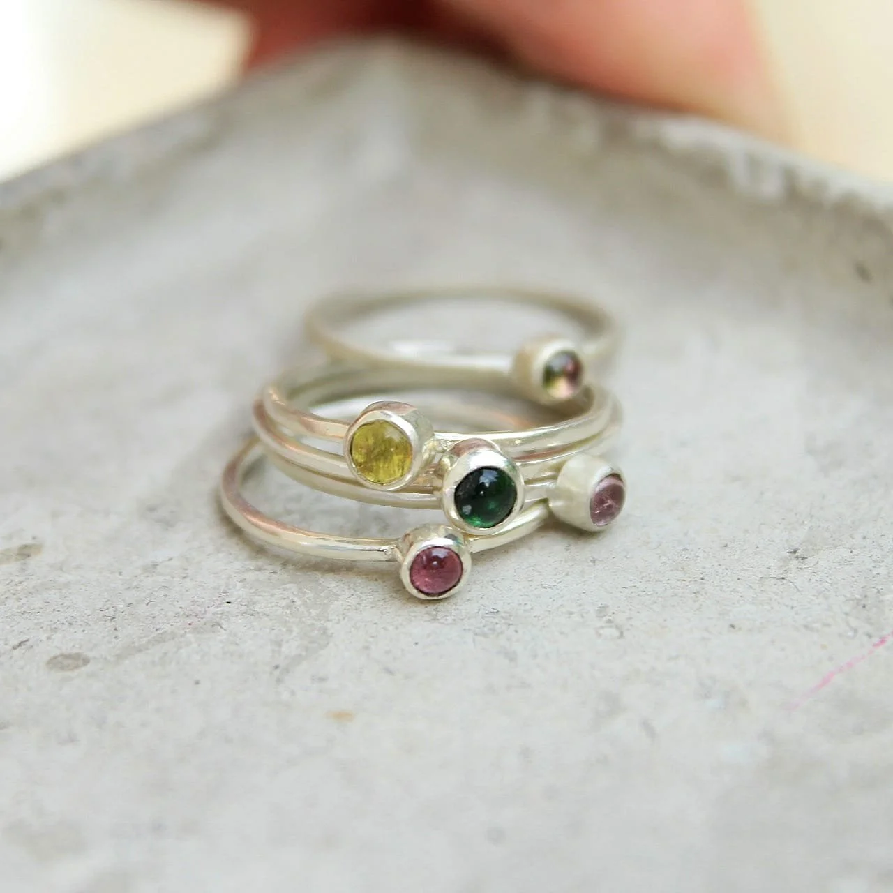 a set of four silver rings with colored gemstones, arranged in ascending order of size, resting on a light gray surface.