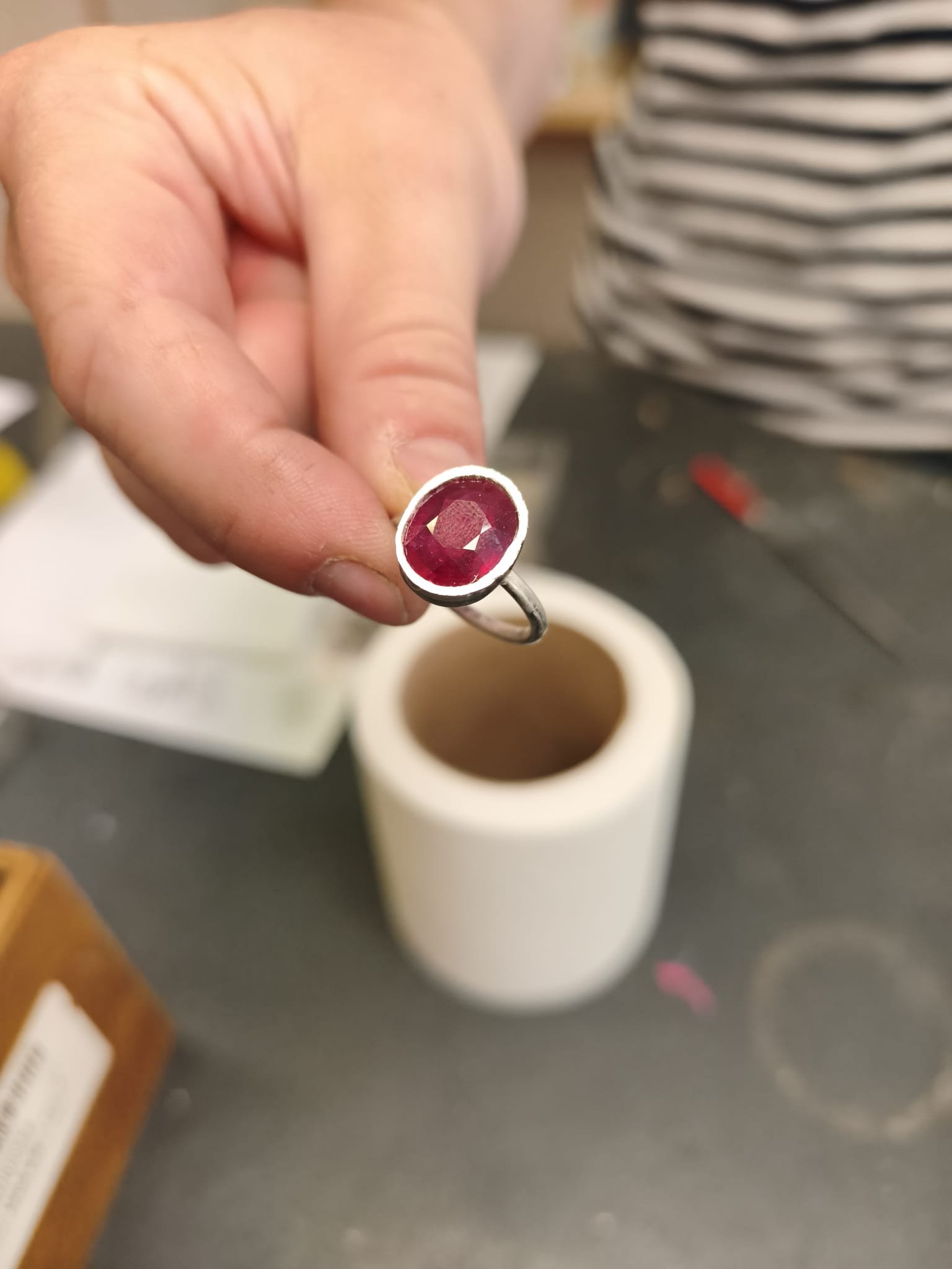 Close-up of a person holding a ring with a large red gemstone, with a white porcelain cup in the background.