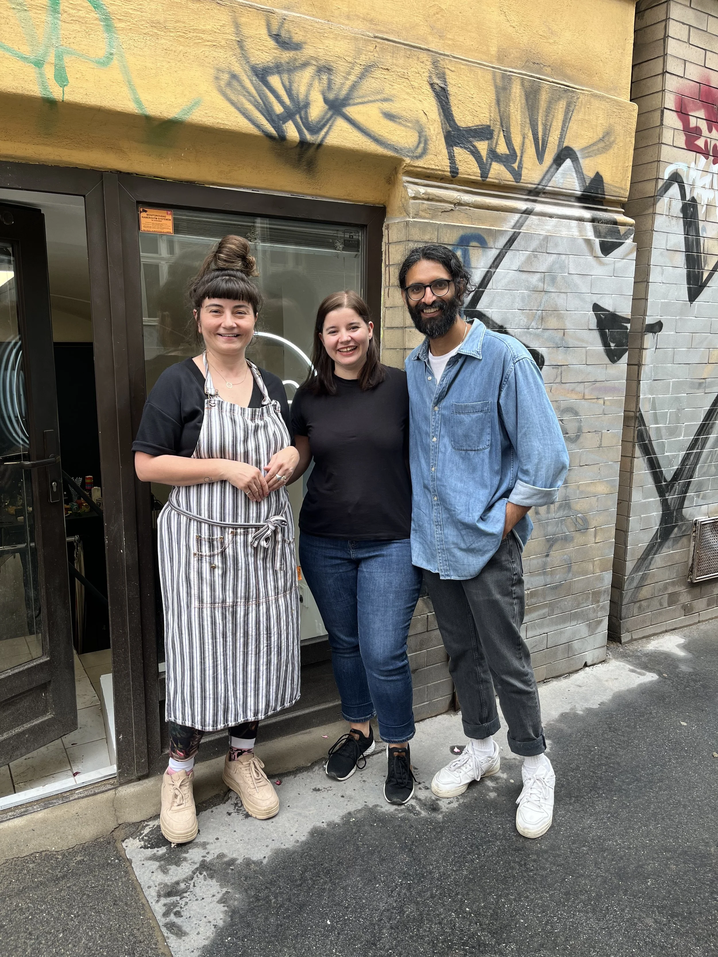 Three people standing outside on a sidewalk in front of a graffiti-covered wall. The woman on the left is wearing a striped apron, black shirt, and beige shoes, with dark hair in a bun. The woman in the middle has brown hair, black shirt, blue jeans,