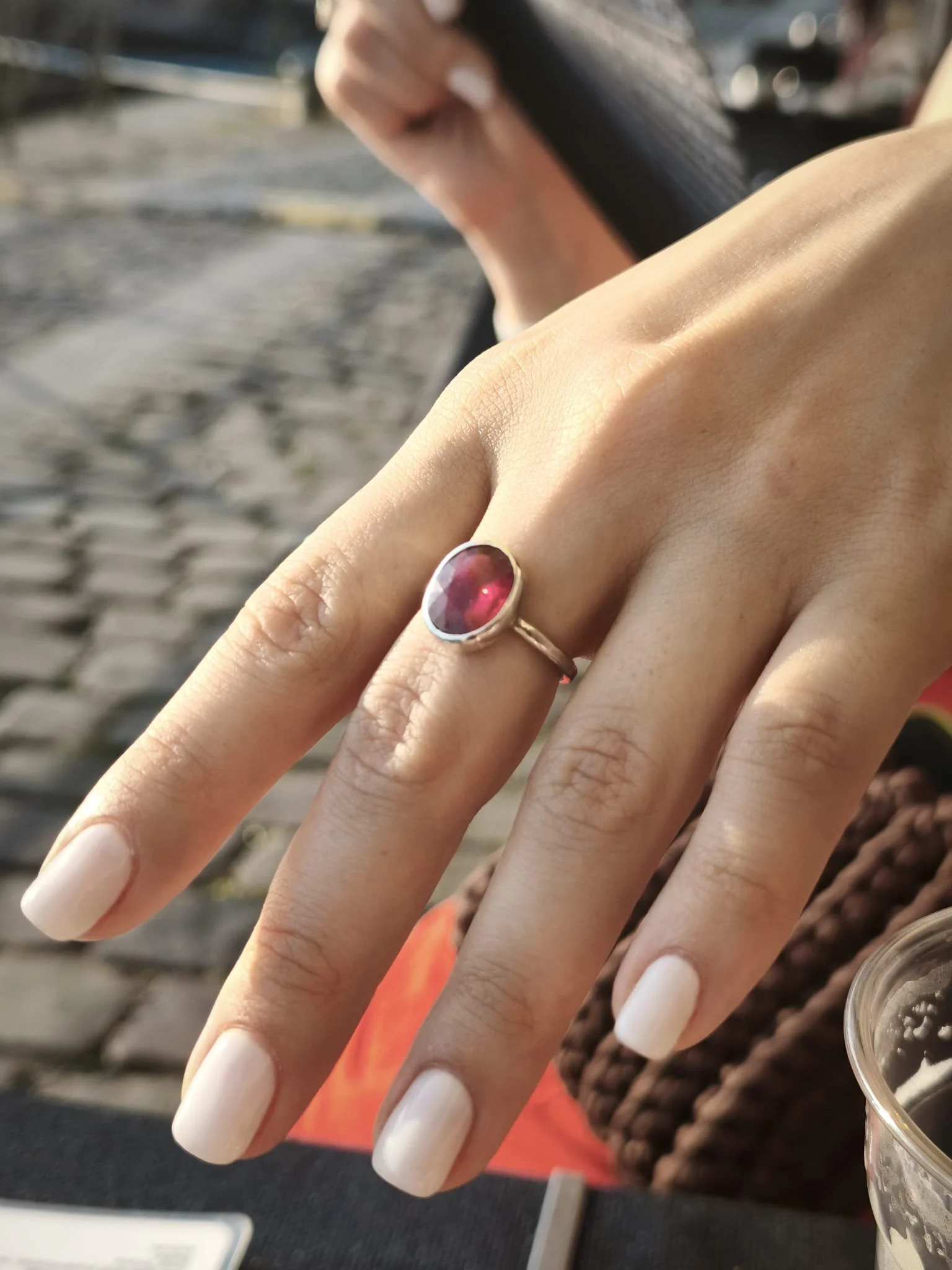 Close-up of a person's hand with a silver ring featuring a red gemstone. The person's nails are manicured with white polish. The background shows a woven brown item and a red rectangular object, with a blurred outdoor setting.